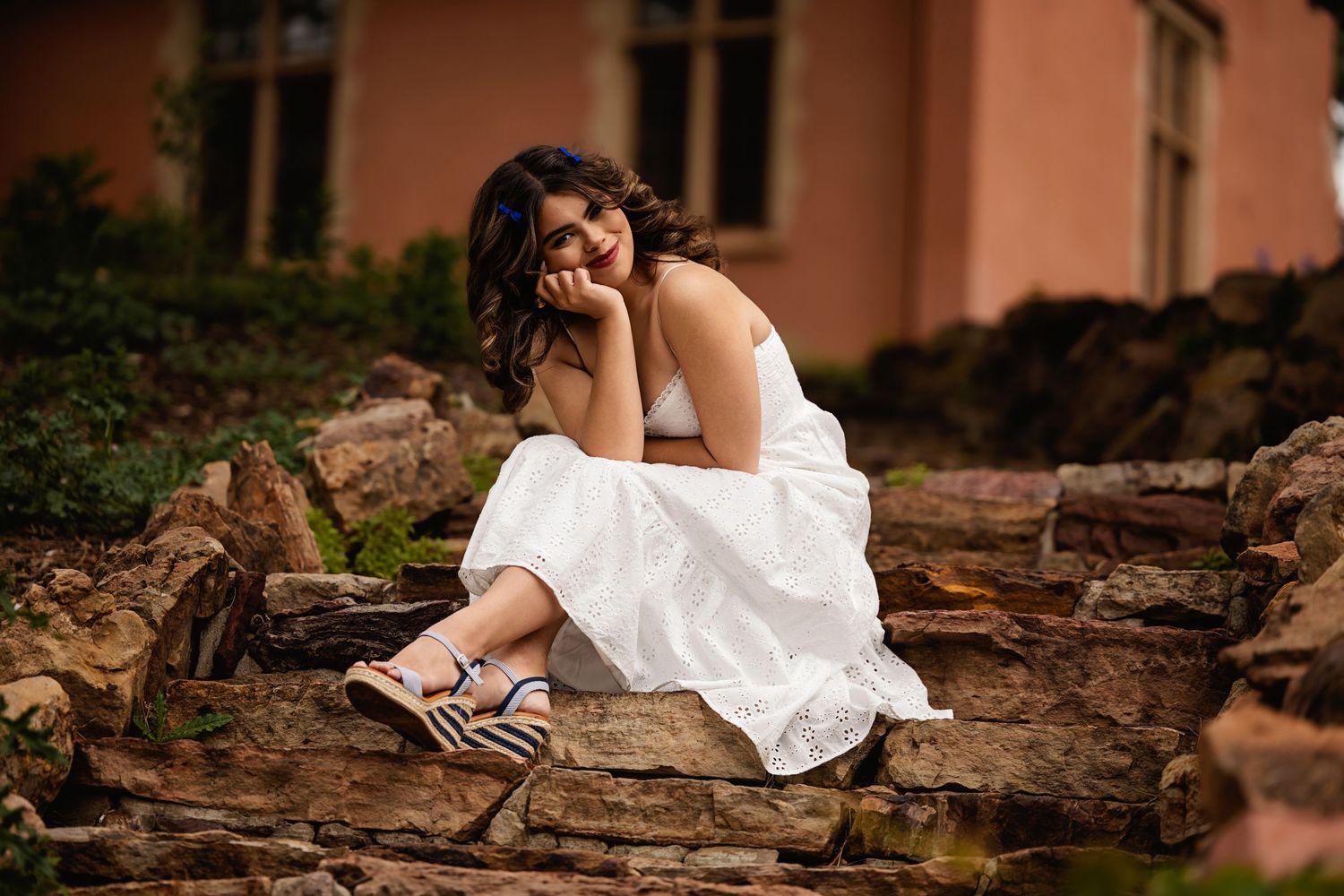 Smiling young woman in white dress sitting on stone steps outdoors, resting head on hand, wearing zebra sandals for Colorado Senior pics