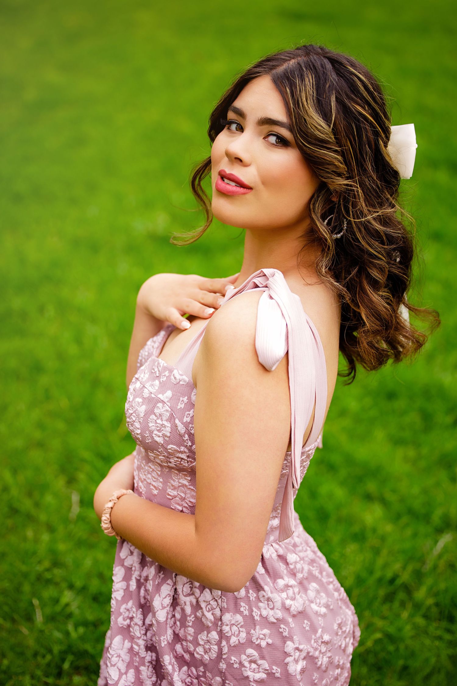 Young woman in pink floral dress with white bow hair accessory posing on lush green grass for Denver Colorado Senior pictures