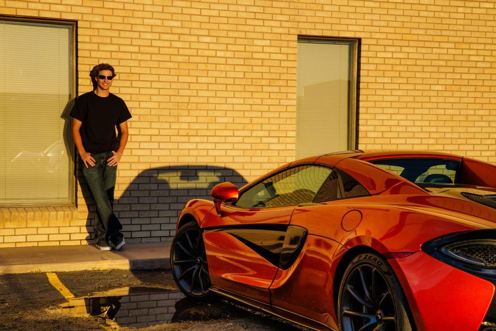 Man in black outfit standing beside a shiny red McLaren sports car against a golden brick wall. Centennial Colorado Senior Portrait