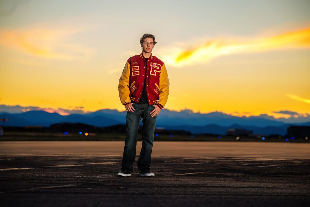 Teen in red and gold varsity jacket stands on dark tarmac under dramatic orange sky. Centennial Colorado Senior Portrait
