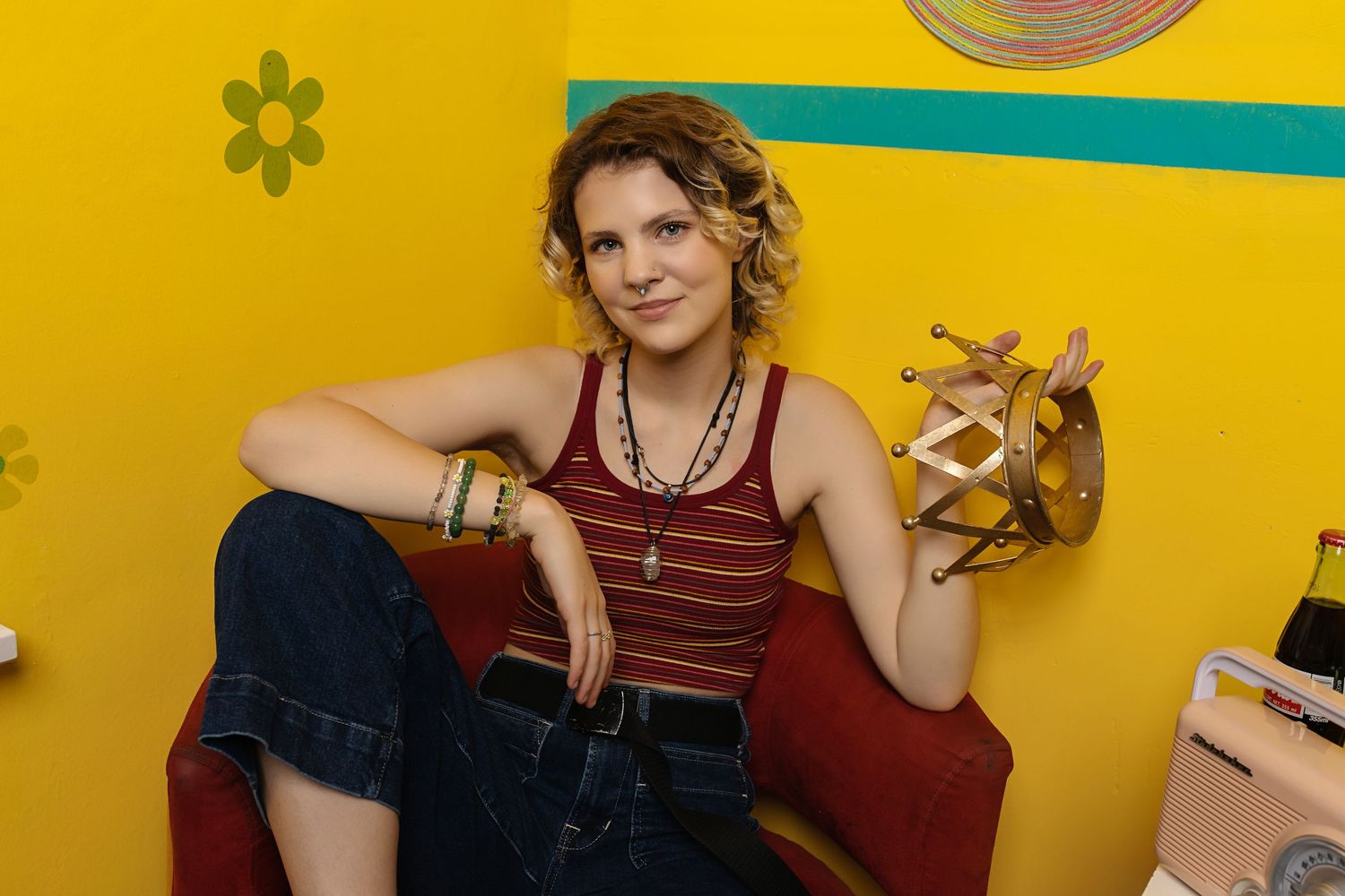 Androgynous teen in red striped tank top and jeans sits relaxed in red chair against bright yellow wall senior photos