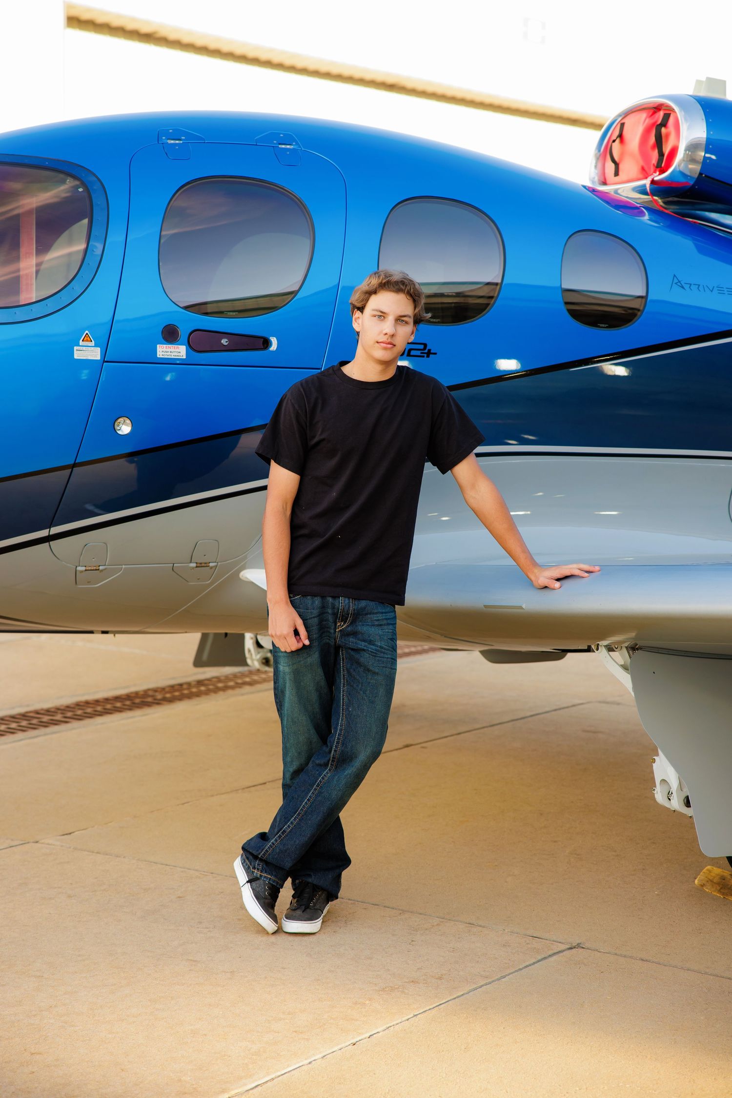 Teen boy in black shirt poses beside a blue Cirrus aircraft on airport pavement.