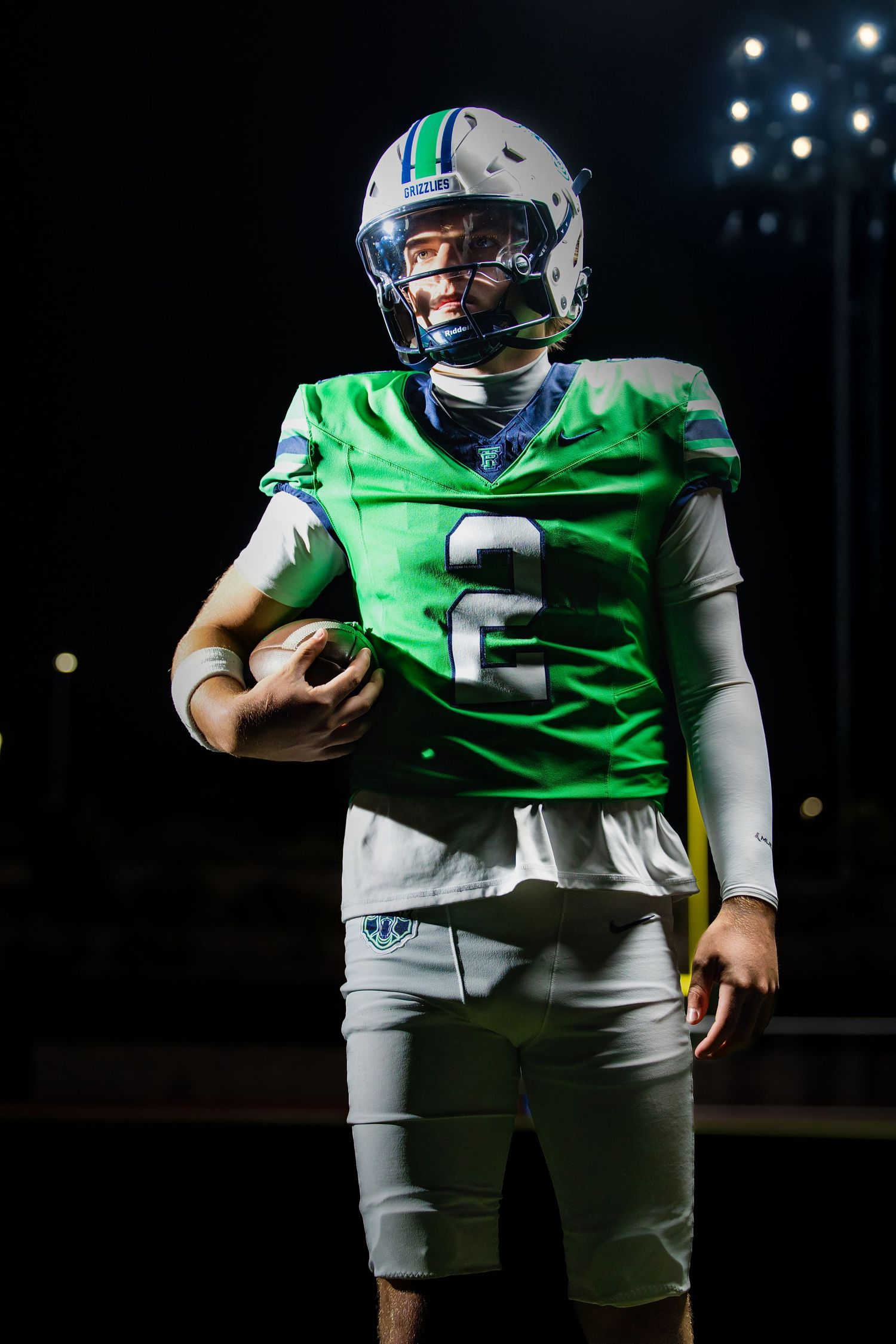 High school football player in green jersey number 2 holding ball under stadium lights at night. Parker Colorado Football Senior Portrait