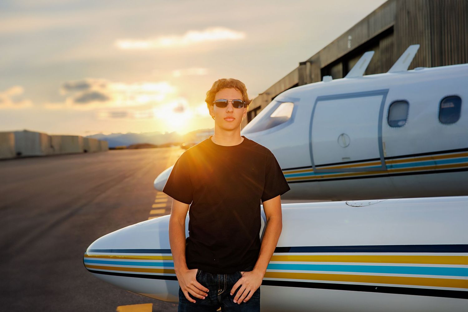 Man in black t-shirt and sunglasses standing by private jet on airport tarmac at sunset.