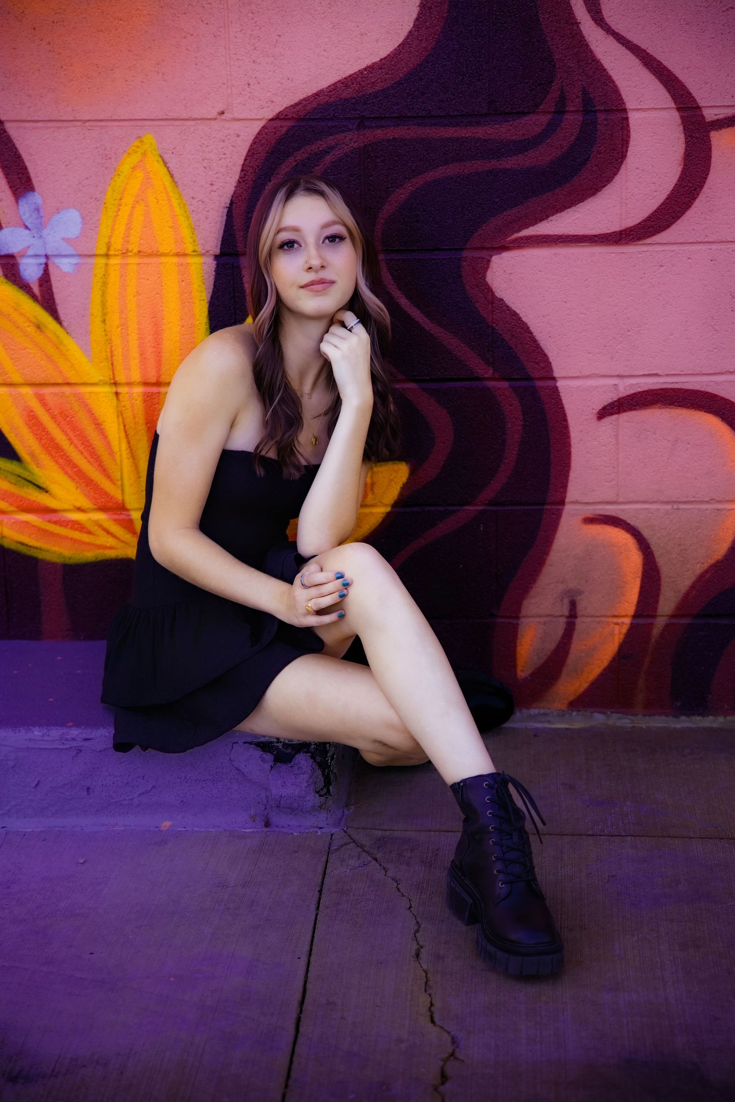Young woman in black dress and combat boots sitting against colorful floral mural wall Urban Downtown Denver CO Senior photos