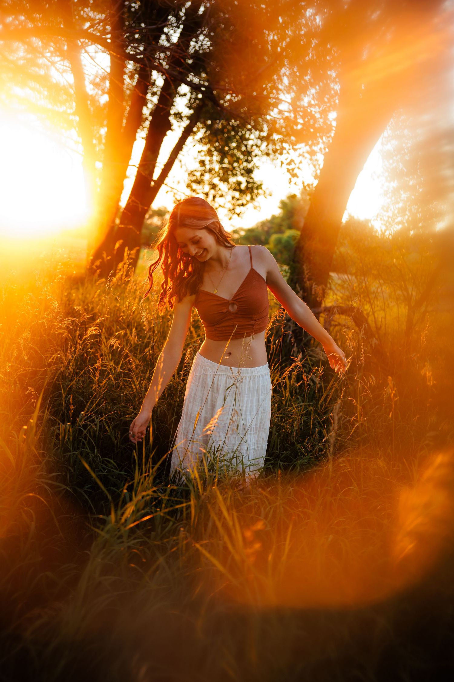 Young woman in crop top and white skirt walking in golden sunset field for Parker CO Senior photos. Film Feel
