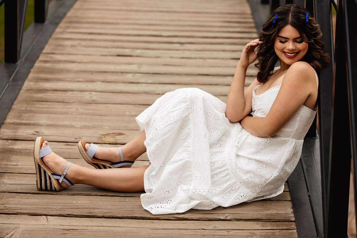 Young woman in white dress sitting against bridge railing wearing striped platform sandals for Parker Colorado Senior pictures