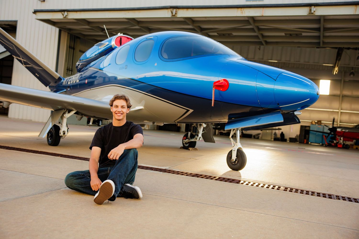 Young man in black shirt sitting on hangar floor in front of a sleek blue private jet.