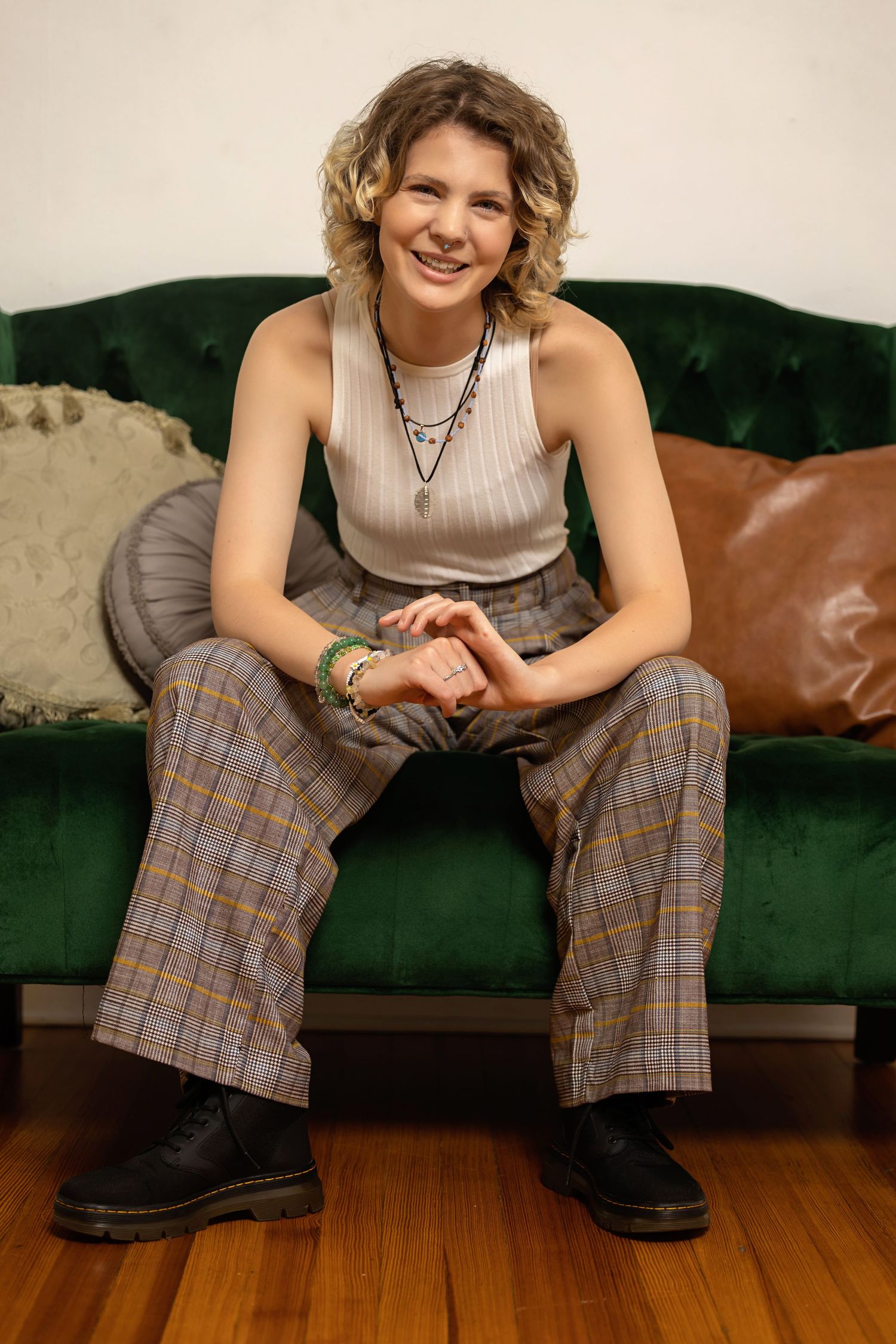 Person  in ribbed tank and plaid wide-leg pants sits smiling on green velvet tufted sofa posing for senior photos downtown Denver studio