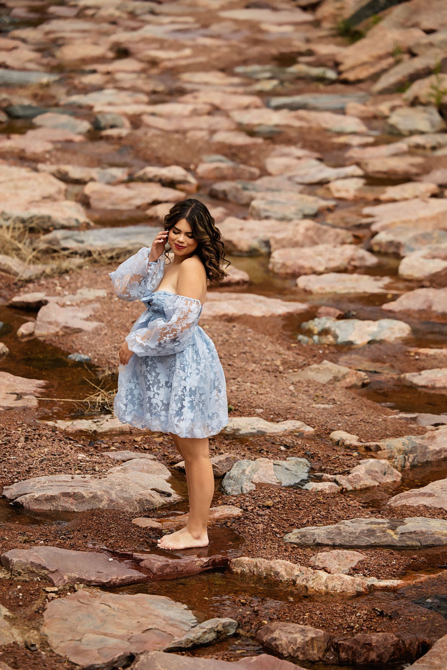 Woman in light blue lace mini dress walking barefoot on rocky creek bed outdoors Arapahoe Country CO High School Senior pictures