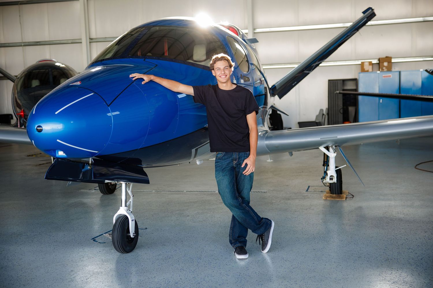Teen boy in black shirt standing beside a blue private aircraft in an airport hangar.