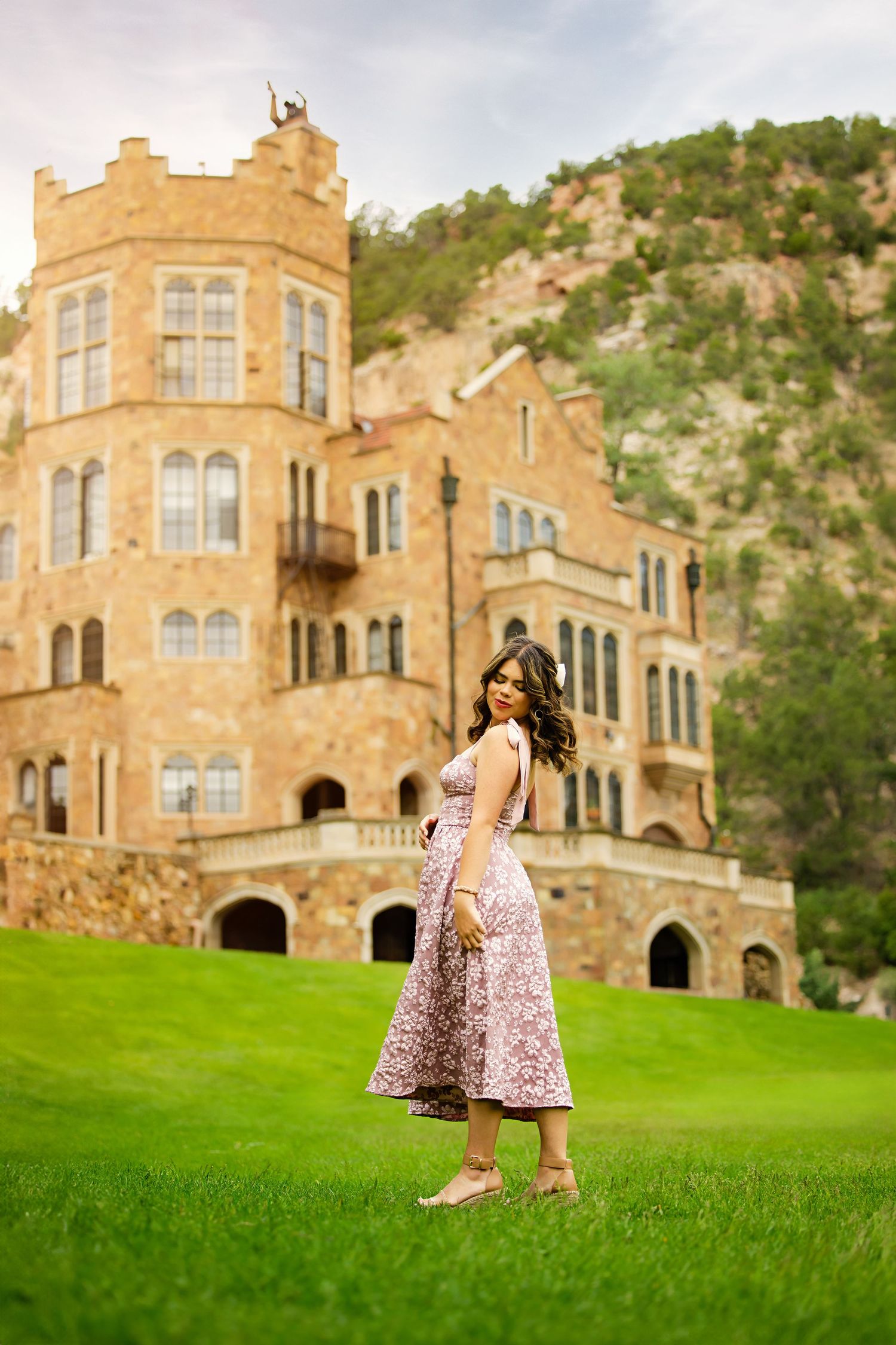 Young woman in pink floral midi dress twirls on green lawn before a grand historic stone castle for Colorado Springs Senior pictures