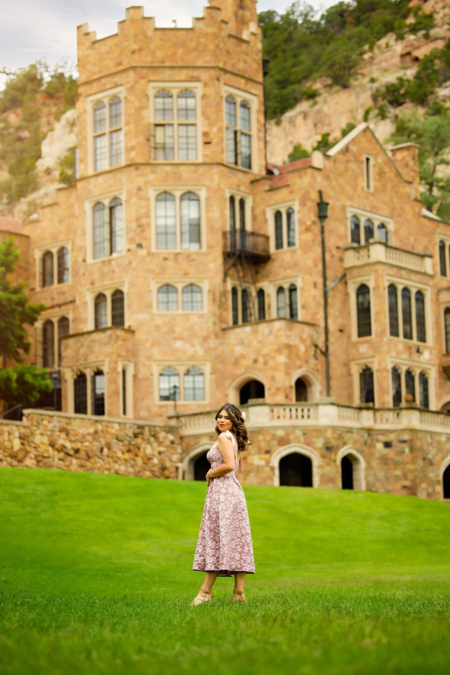 Young woman in pink floral dress posing on green lawn in front of large stone castle for Parker Colorado Senior pictures