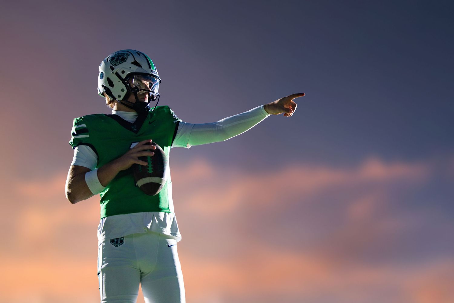 Football quarterback in green uniform pointing downfield at sunset, wearing helmet and holding football.