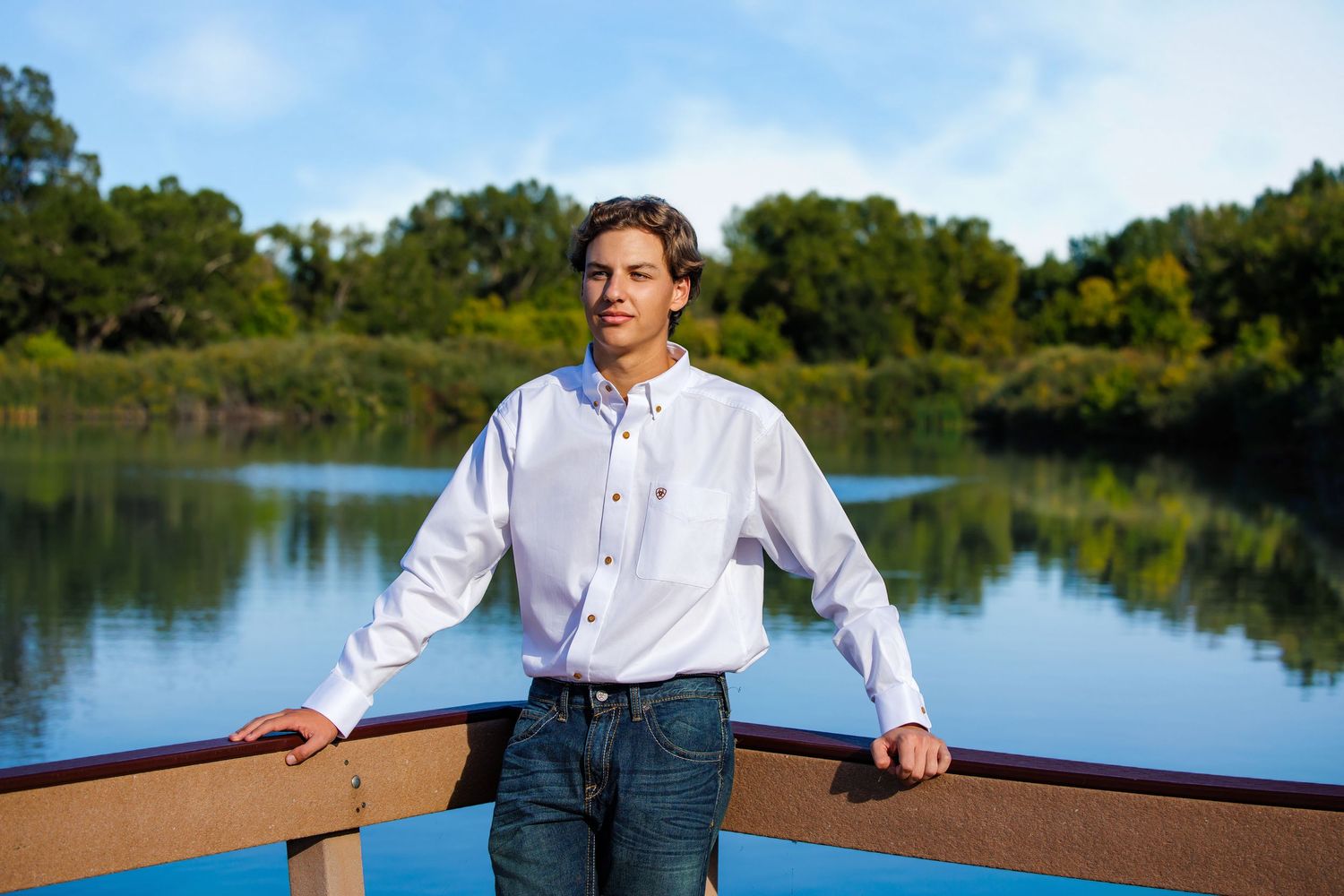 Young man in white shirt leaning on wooden railing with scenic lake and trees behind him. Highlands Ranch Colorado Senior Portrait