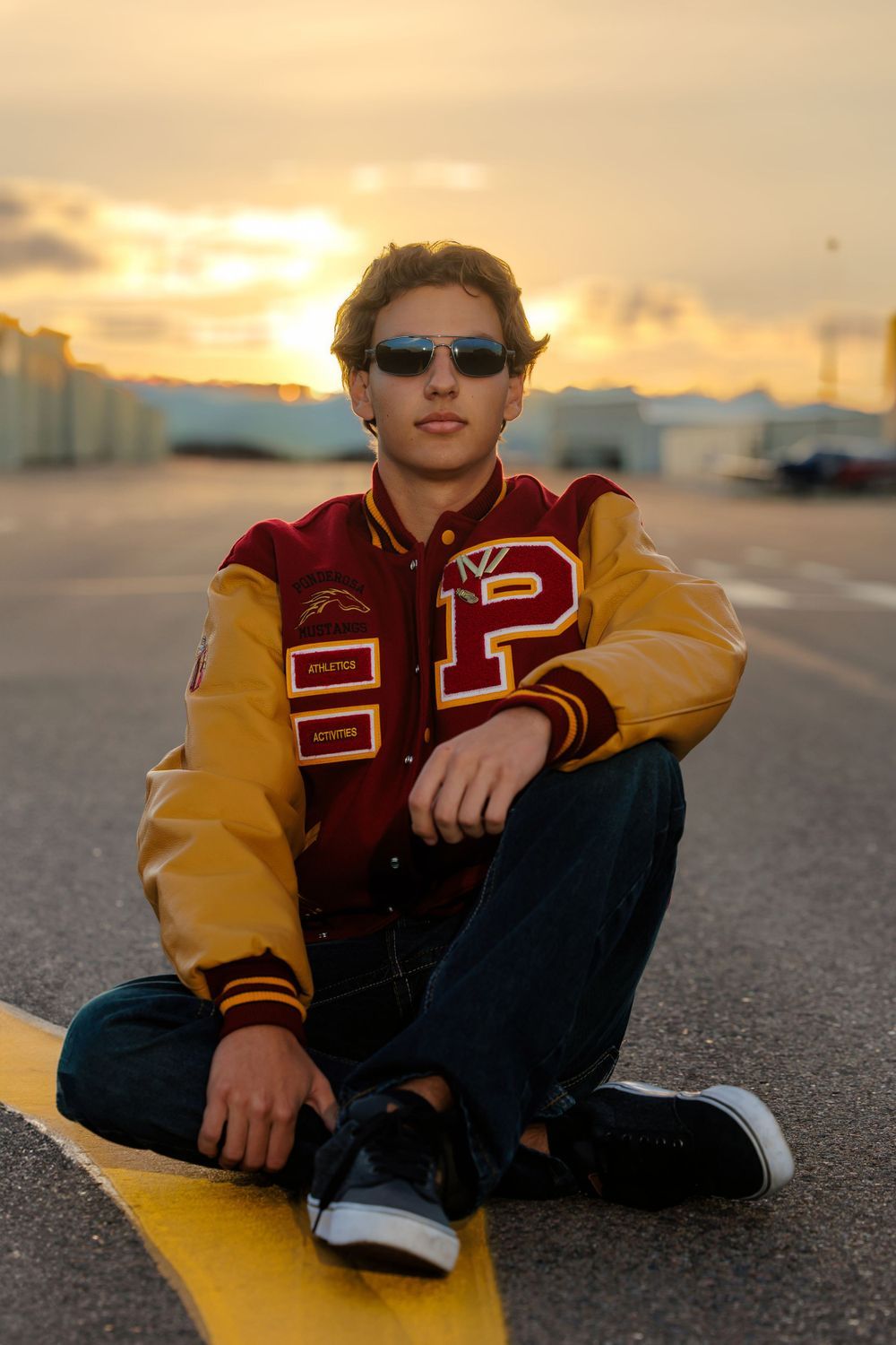 Teen boy in red varsity jacket and sunglasses sits on yellow runway line at golden sunset. Centennial Colorado Senior Portrait
