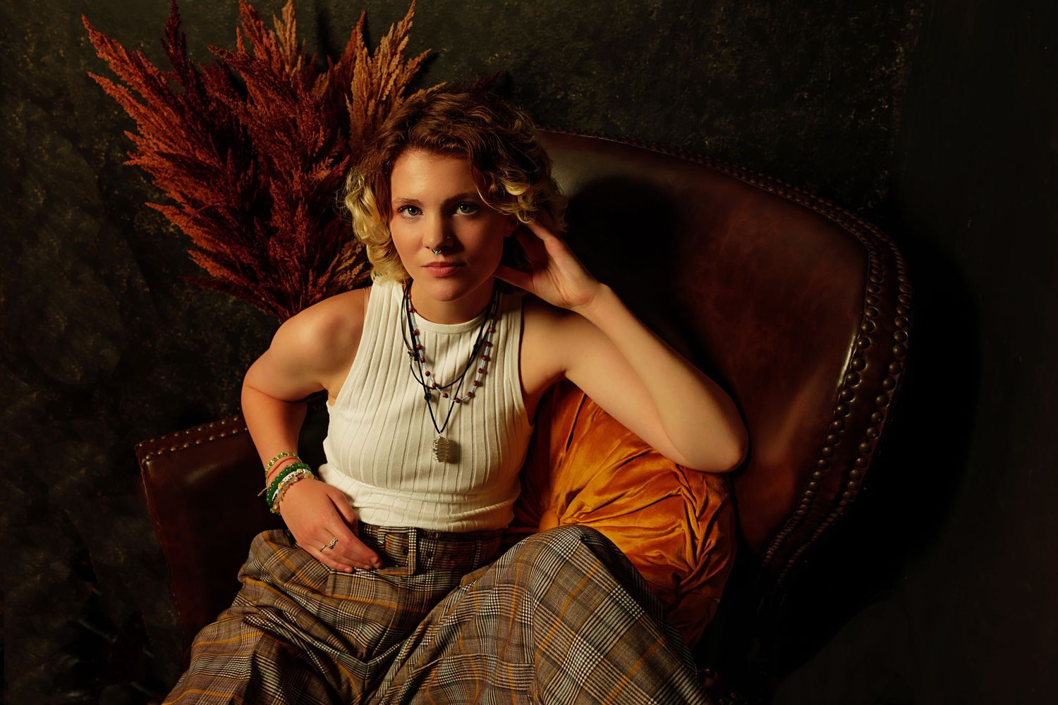 Teen in white tank top and plaid pants relaxes in brown leather chair with autumn dried floral decor.