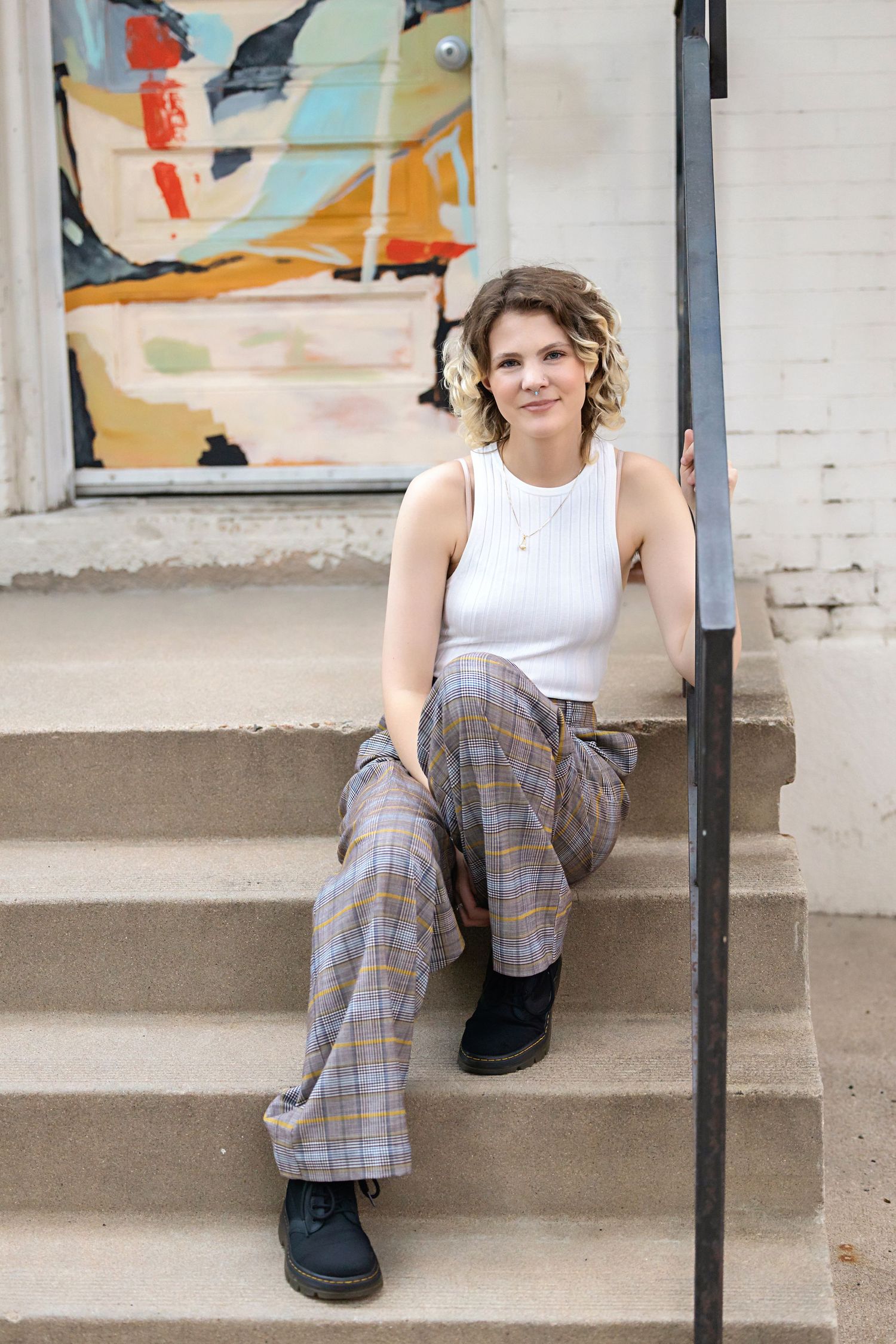 Androgynous person in white tank top and plaid pants sitting on outdoor steps near colorful abstract mural. Parker Senior Photos