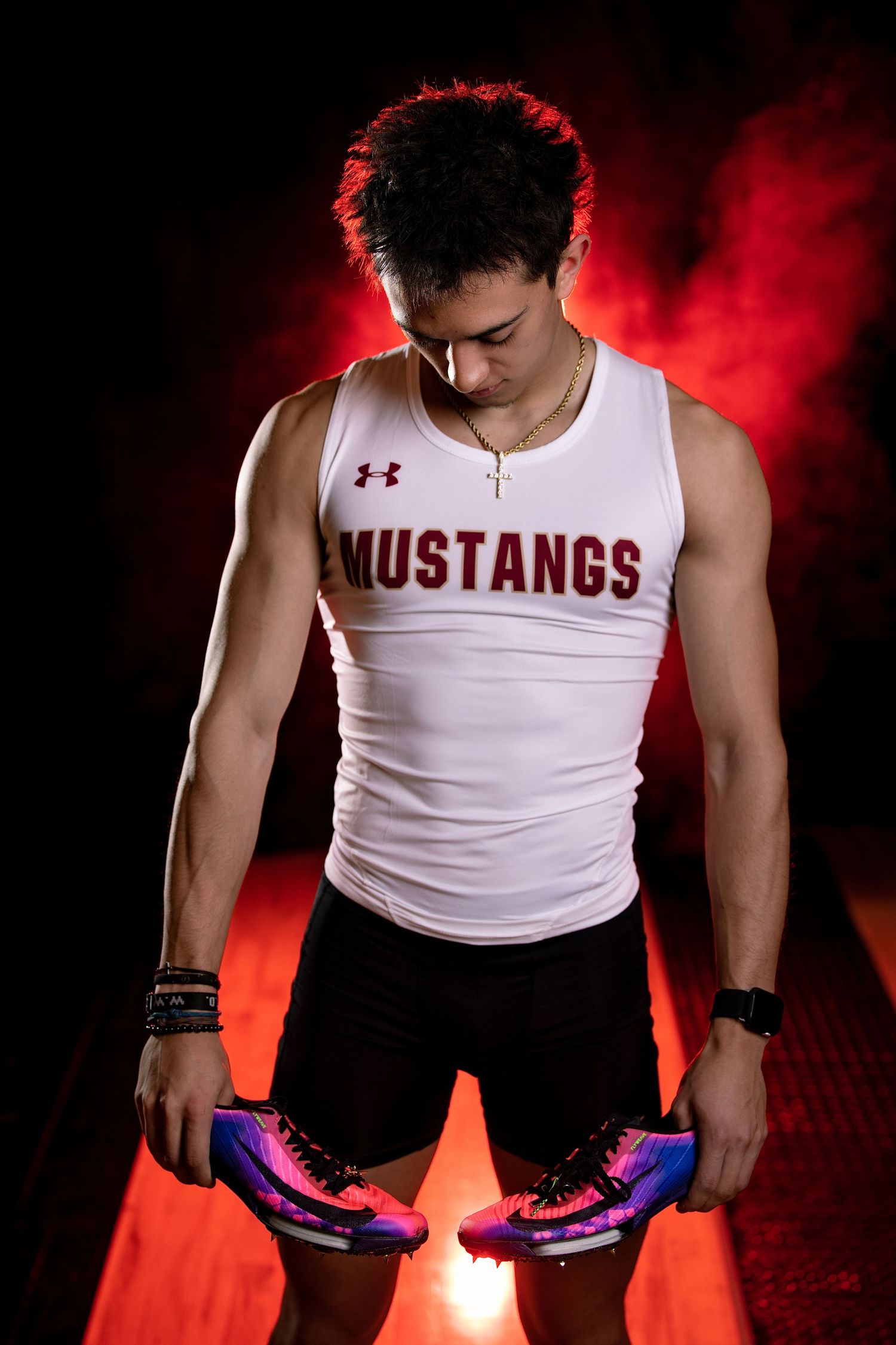 Athletic teen in Mustangs track uniform holding colorful running shoes under red dramatic lighting. Athletic portraits Parker Colorado