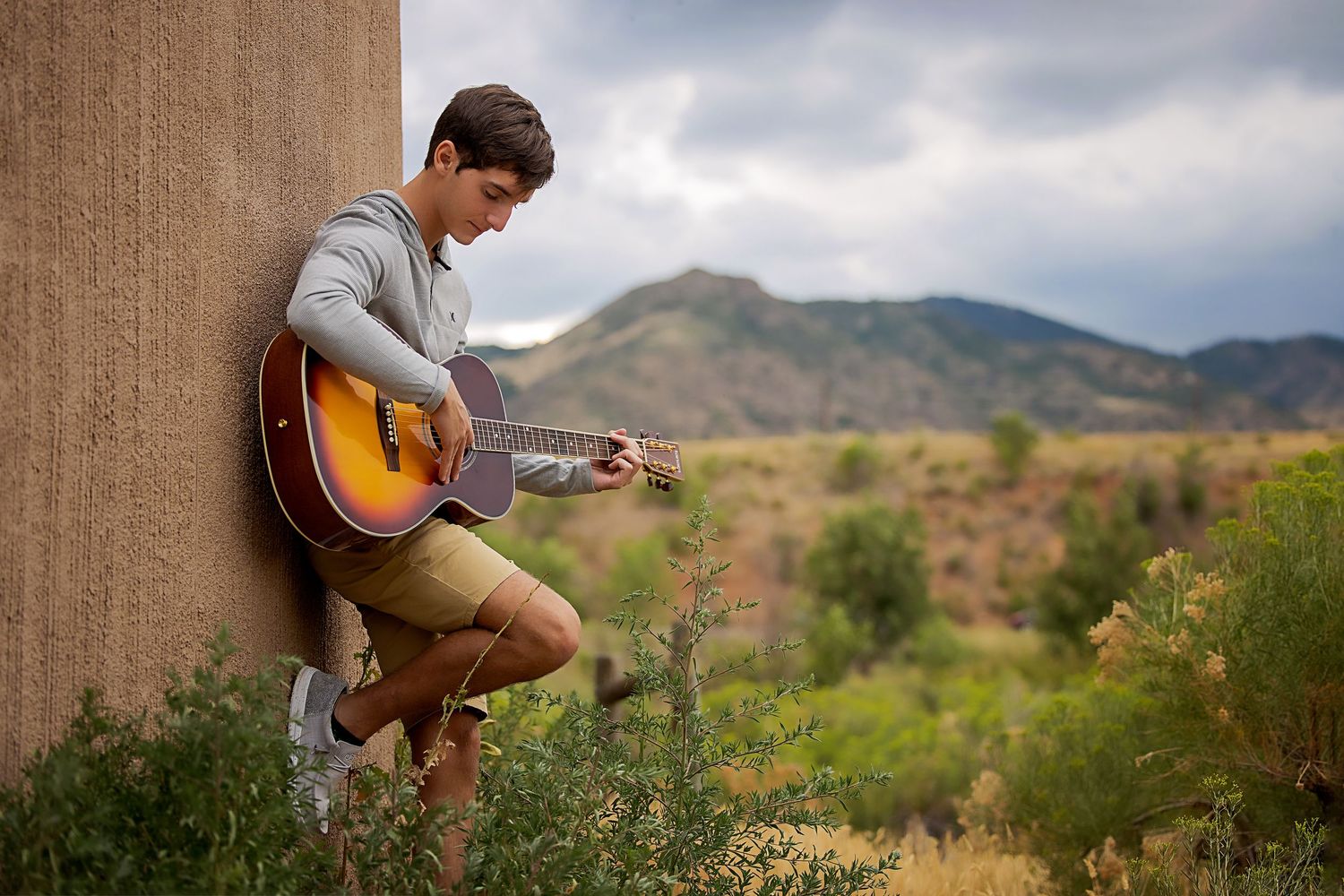 CO High School man playing acoustic guitar outdoors, leaning against a wall with scenic mountain landscape in background Highlands Ranch CO