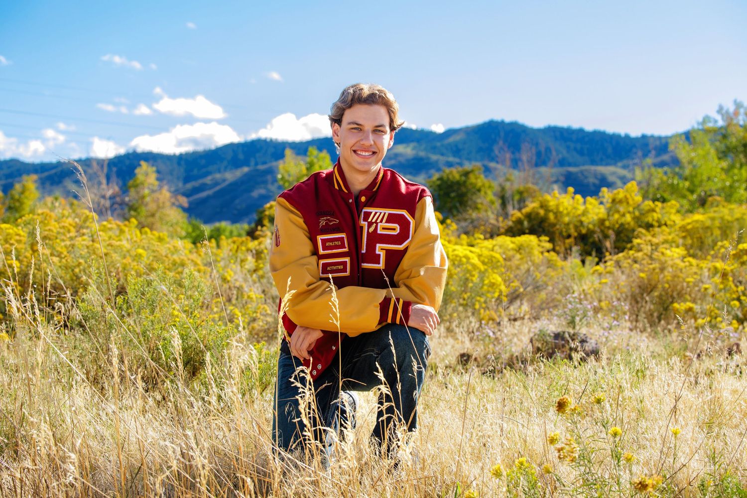 Teen boy in red gold varsity jacket kneeling in golden field for senior portraits. Highlands Ranch Colorado Senior Portrait