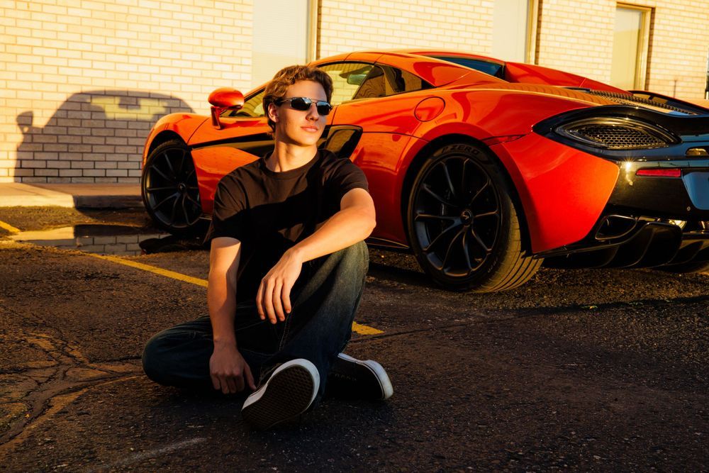 Teen boy in black shirt sits on pavement in front of a vibrant orange McLaren sports car. Centennial Colorado Senior Portrait