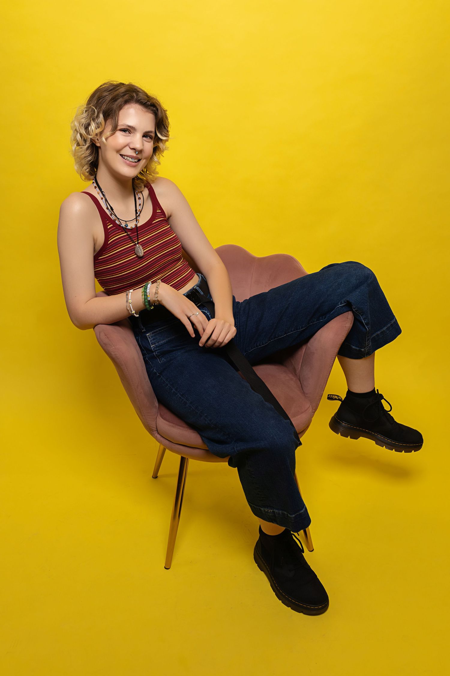 Androgynous person in striped red tank and dark jeans sits in pink chair against bright yellow background.