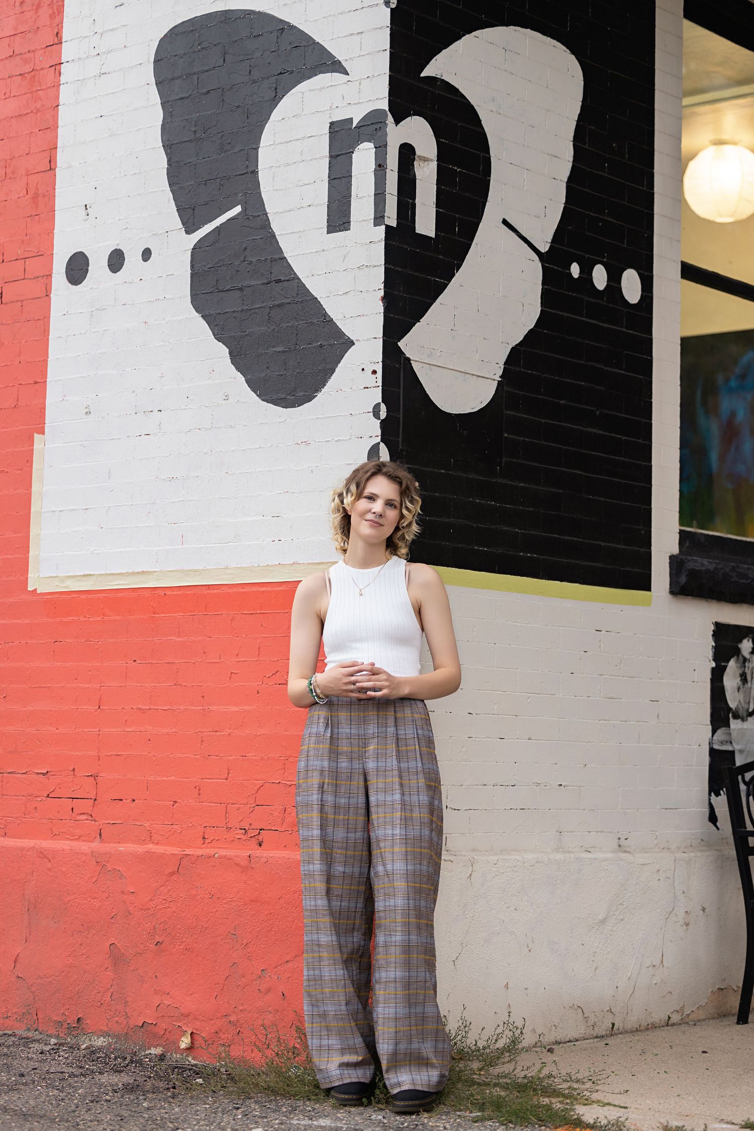 Androgynous teen in white tank and plaid wide-leg pants standing beside bold black and red mural for high school senior photos