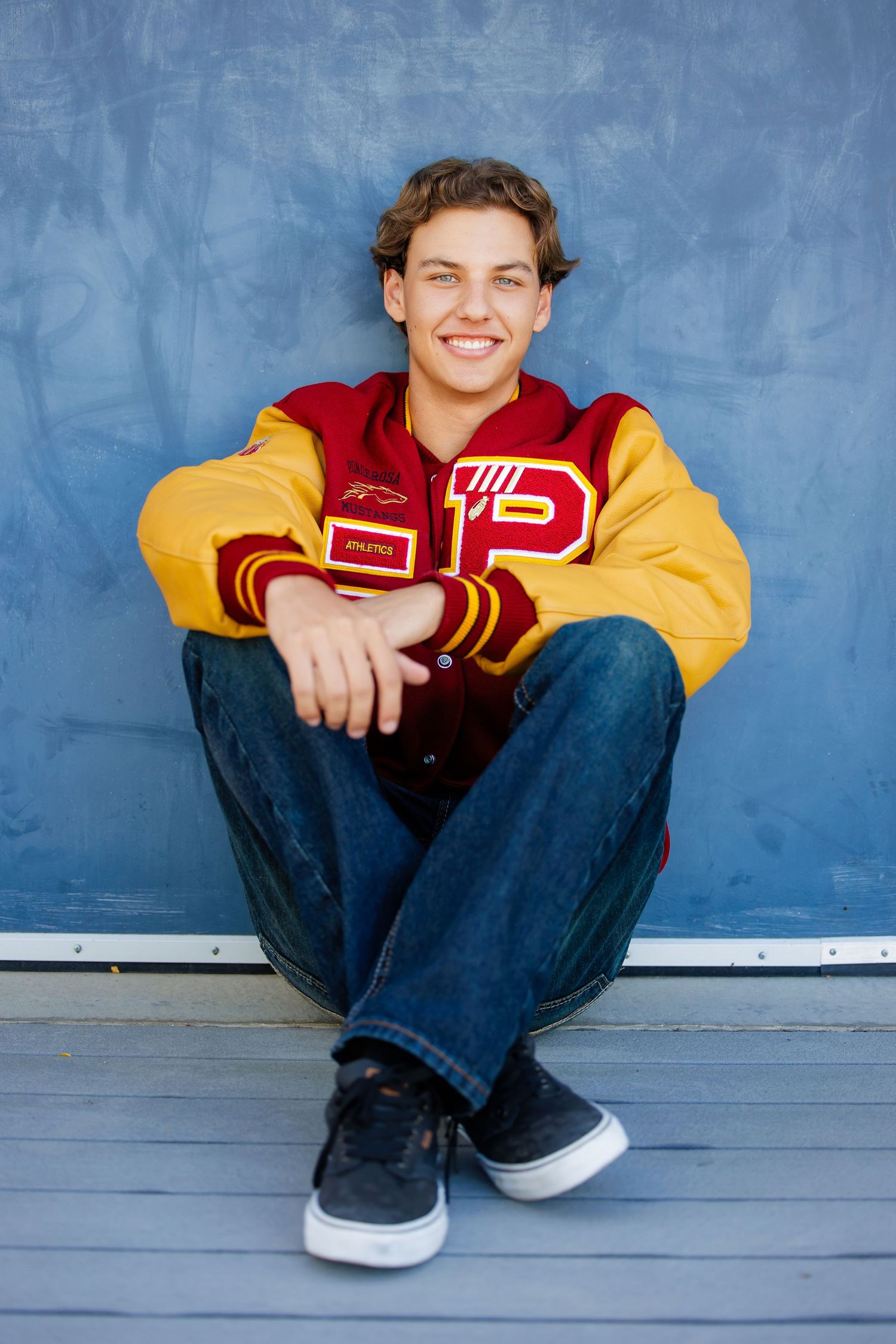 Happy teen boy in varsity jacket sitting cross-legged on ground smiling for senior photos.  Aurora Colorado Senior Portrait