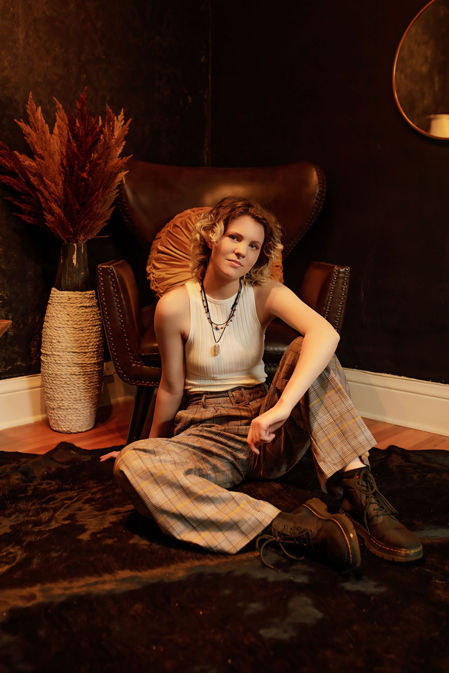 Androgynous person in plaid wide-leg pants and white tank sits on floor beside leather chair in moody dark room.