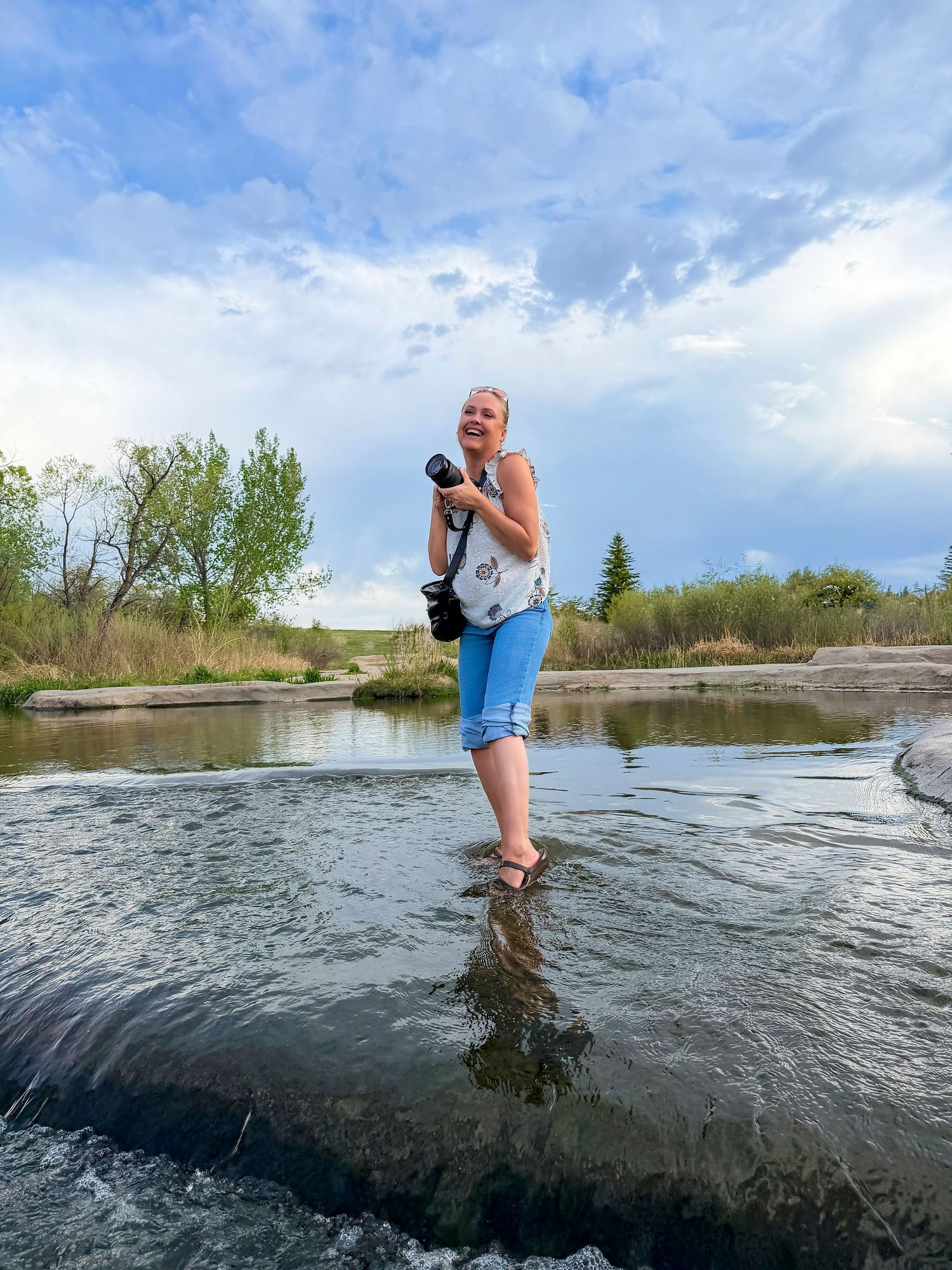 Woman photographer standing in shallow river holding camera on cloudy day.