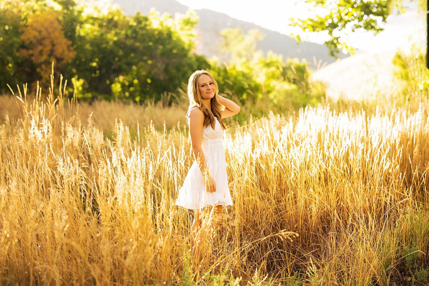Young woman in white dress standing in golden wheat field with sunlit trees and mountains in background.
