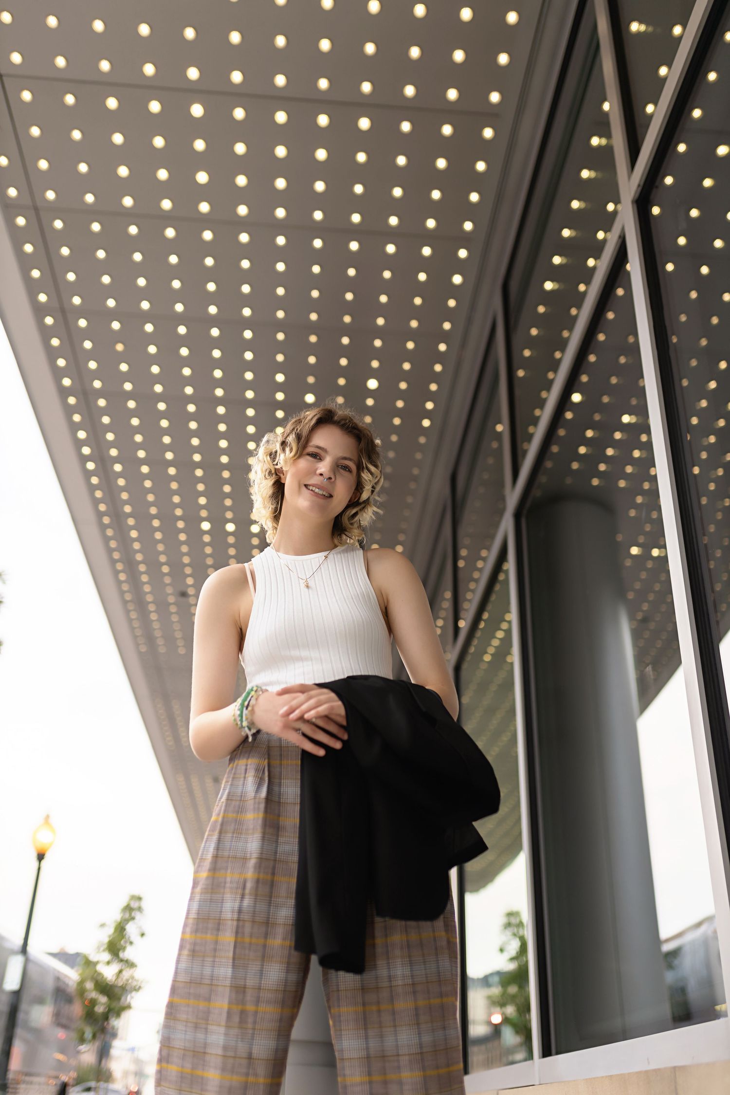 Smiling teen in white top and plaid pants holds black blazer under decorative marquee lights. Denver LQBTQ ally senior photographer