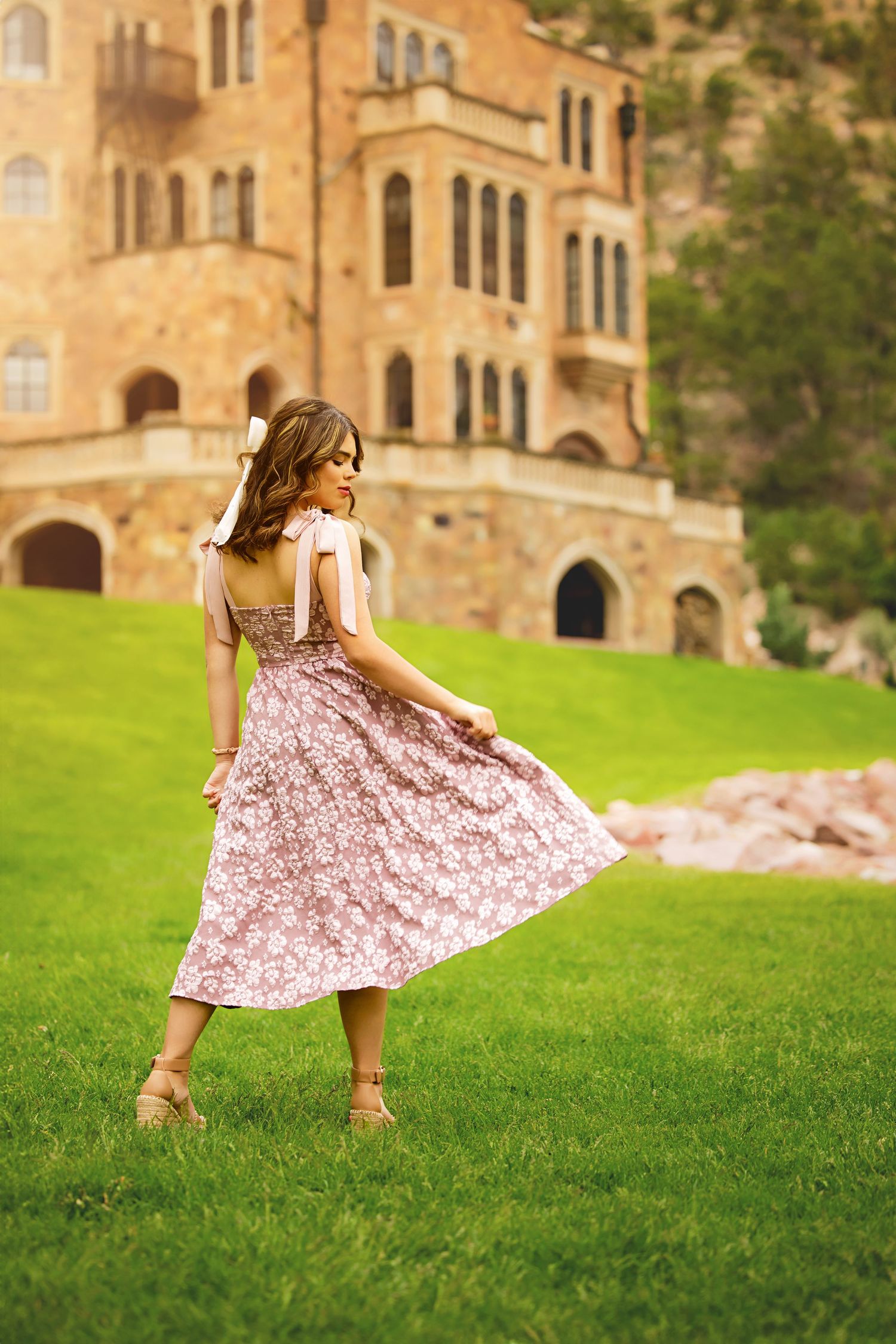 Teen in floral pink midi dress twirling on green lawn in front of historic stone castle building posing for Colorado Senior photos