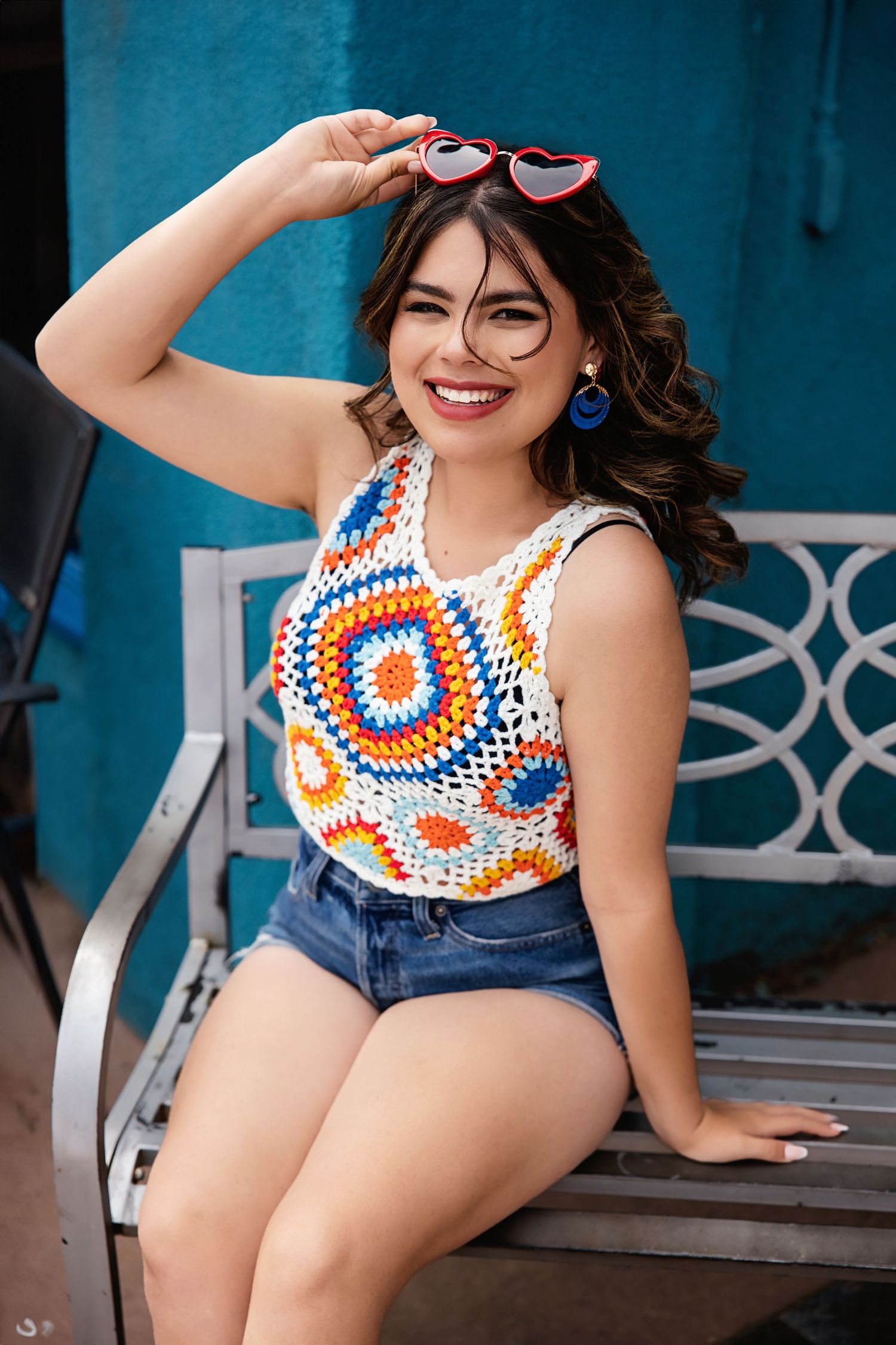 Smiling teen in colorful crochet top and denim shorts sitting on white bench with heart sunglasses for High School Senior pictures