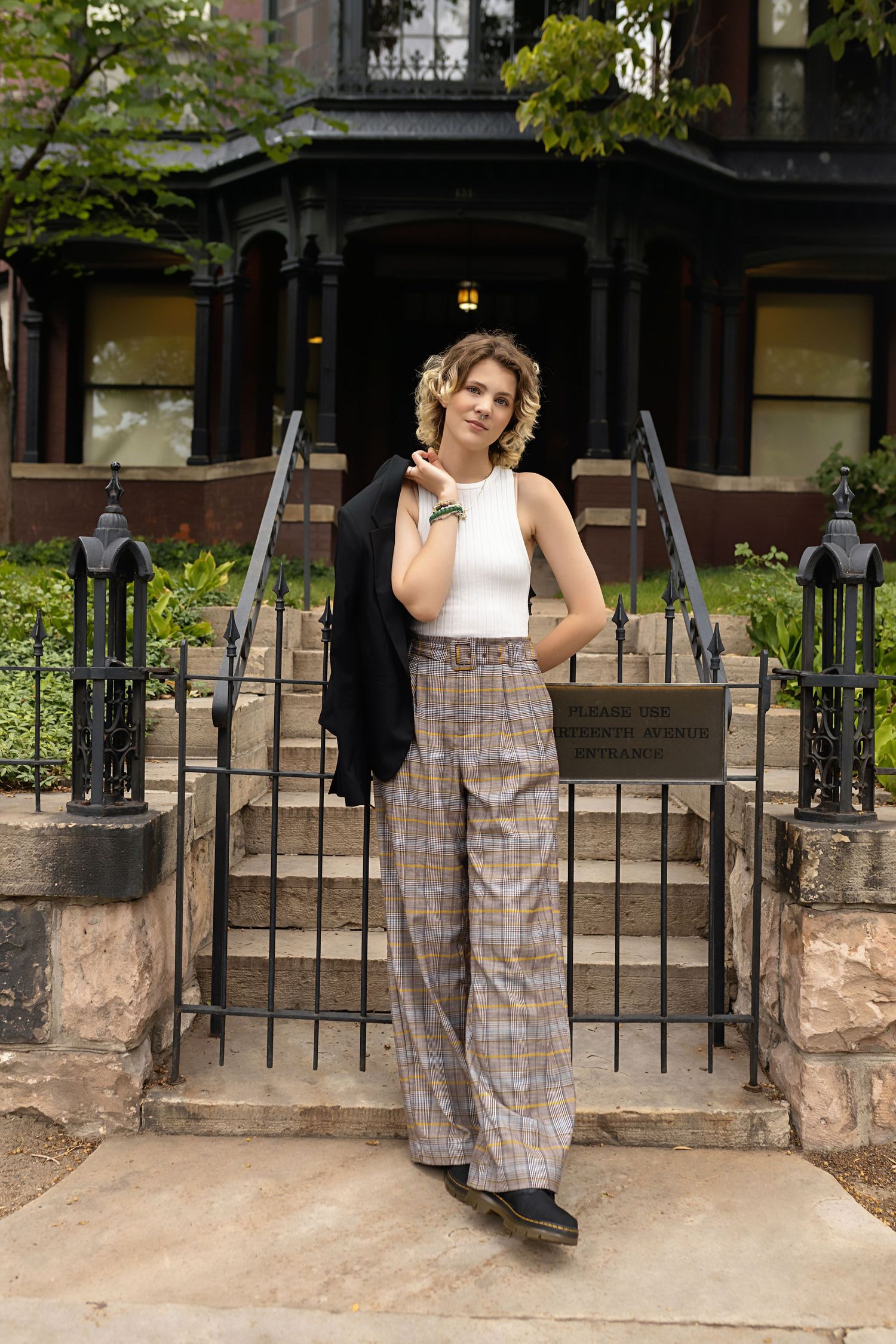 Fashionable teen in plaid wide-leg pants carries black blazer while posing at historic building steps. Denver urban senior photos