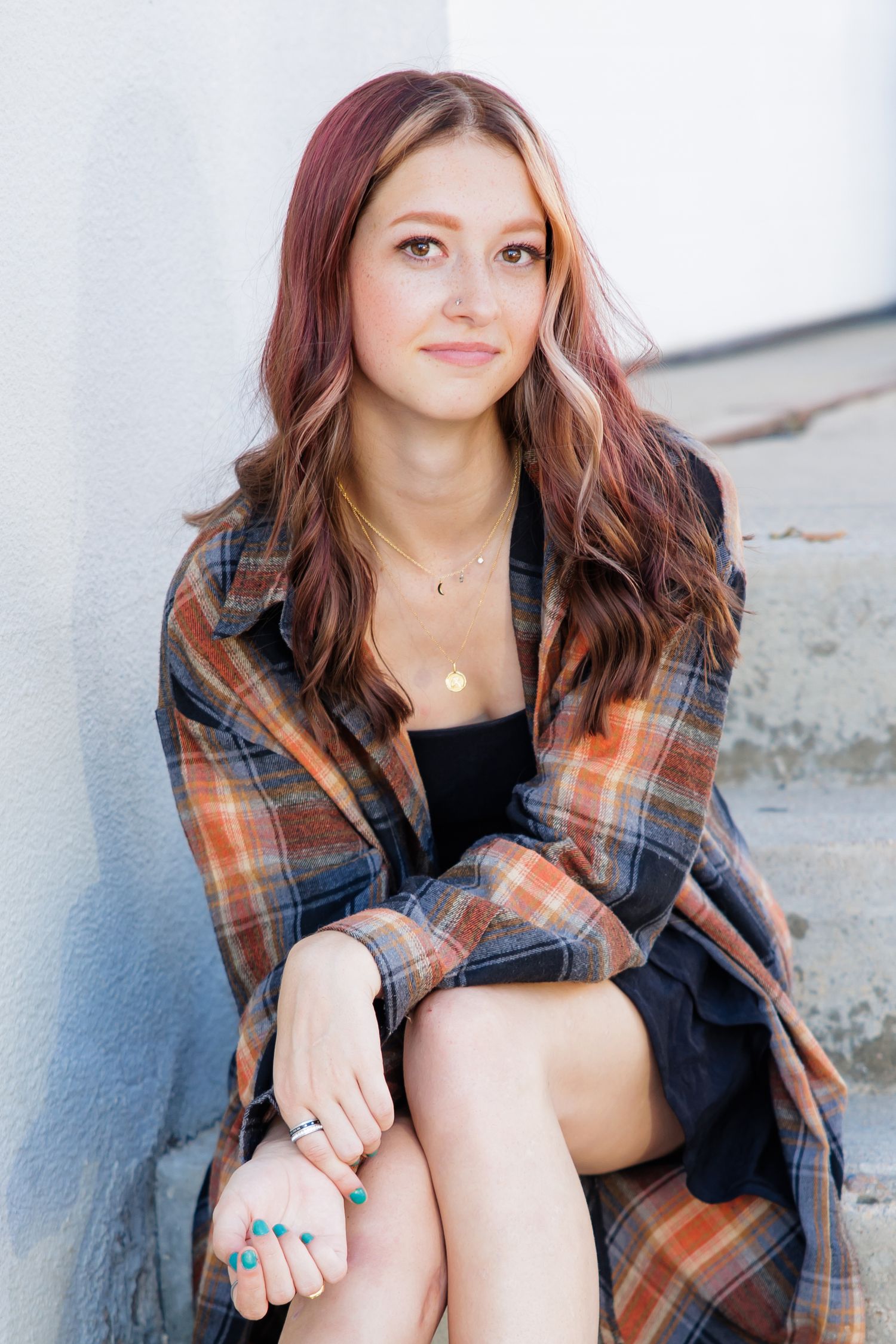 Young woman with auburn hair wearing plaid coat sitting on steps against a white wall for Urban Parker Colorado Senior pictures