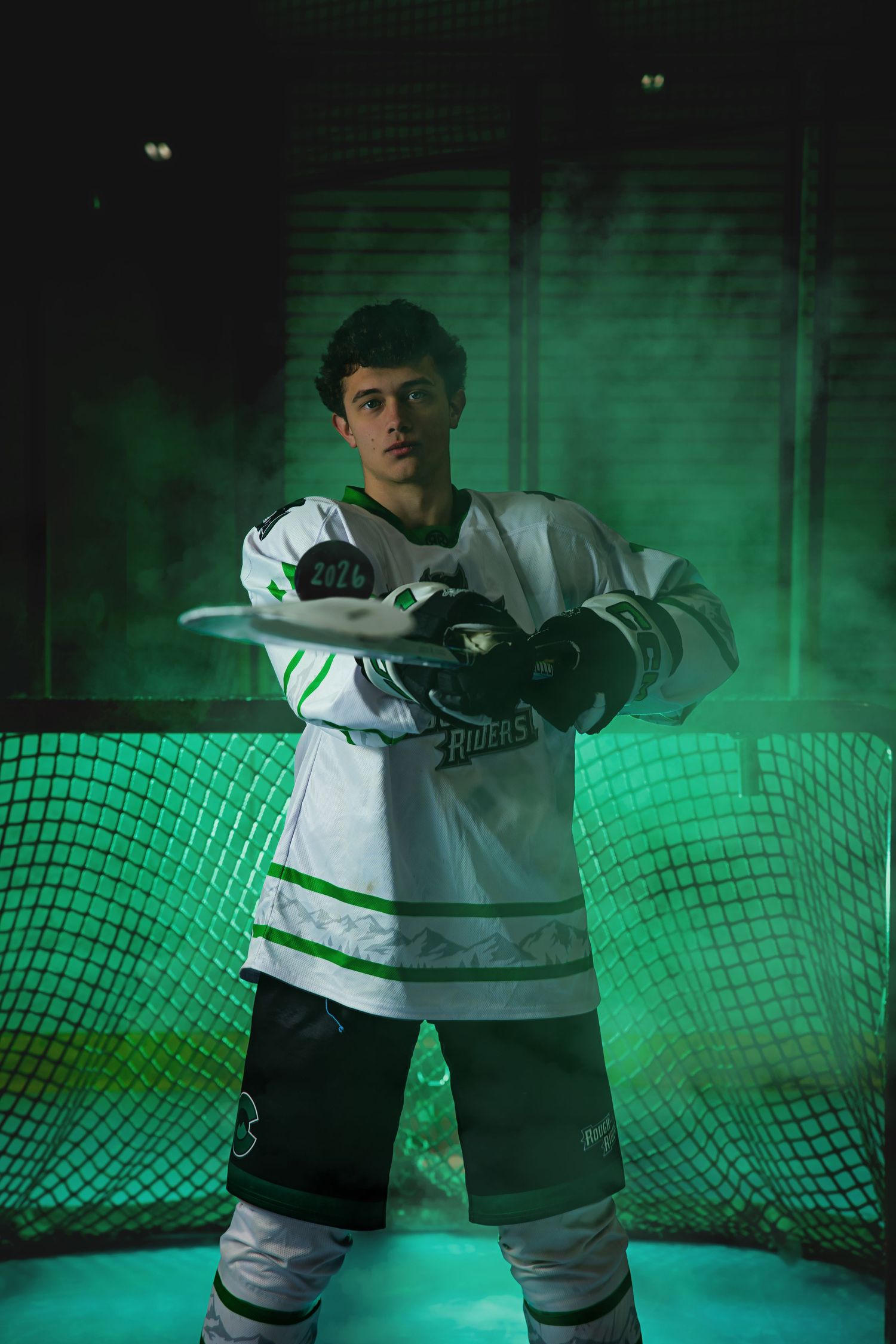Male hockey player in white Riders jersey posing dramatically with stick in green smoky arena.  Colorado Hockey Senior Portrait