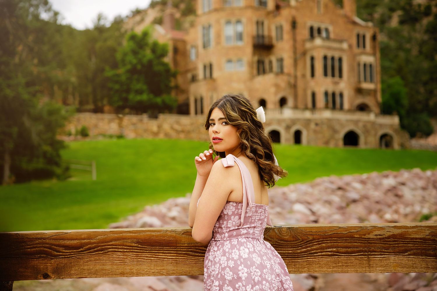 Young woman in floral pink dress posing near fence with historic stone castle building in background for Parker Colorado Senior pictures