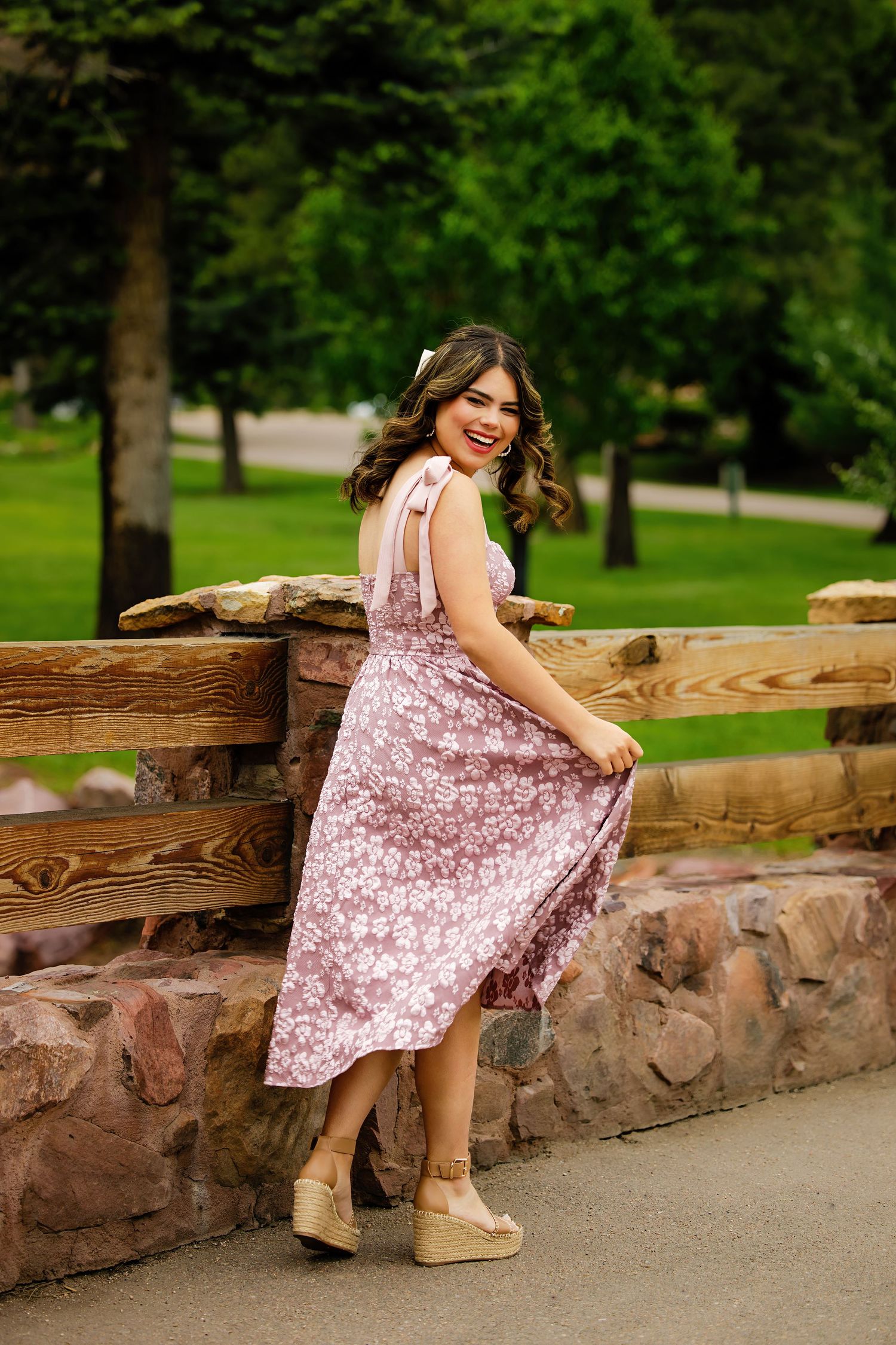 Young woman in pink floral dress lifts skirt while leaning on rustic wooden fence in green park for Aurora Colorado Senior pictures