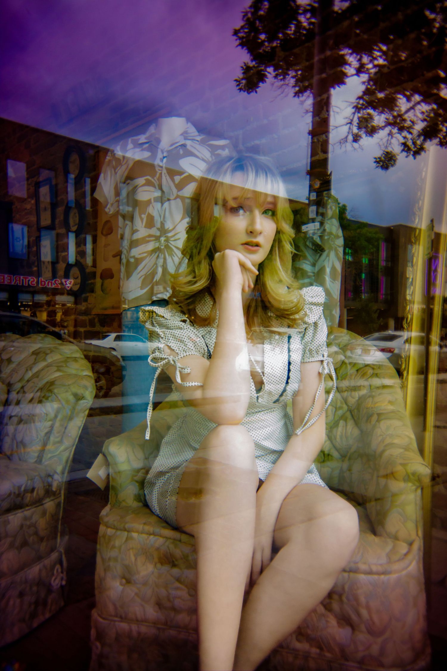 A blonde mannequin in a white dress sits in a floral chair, visible through a reflective store window.