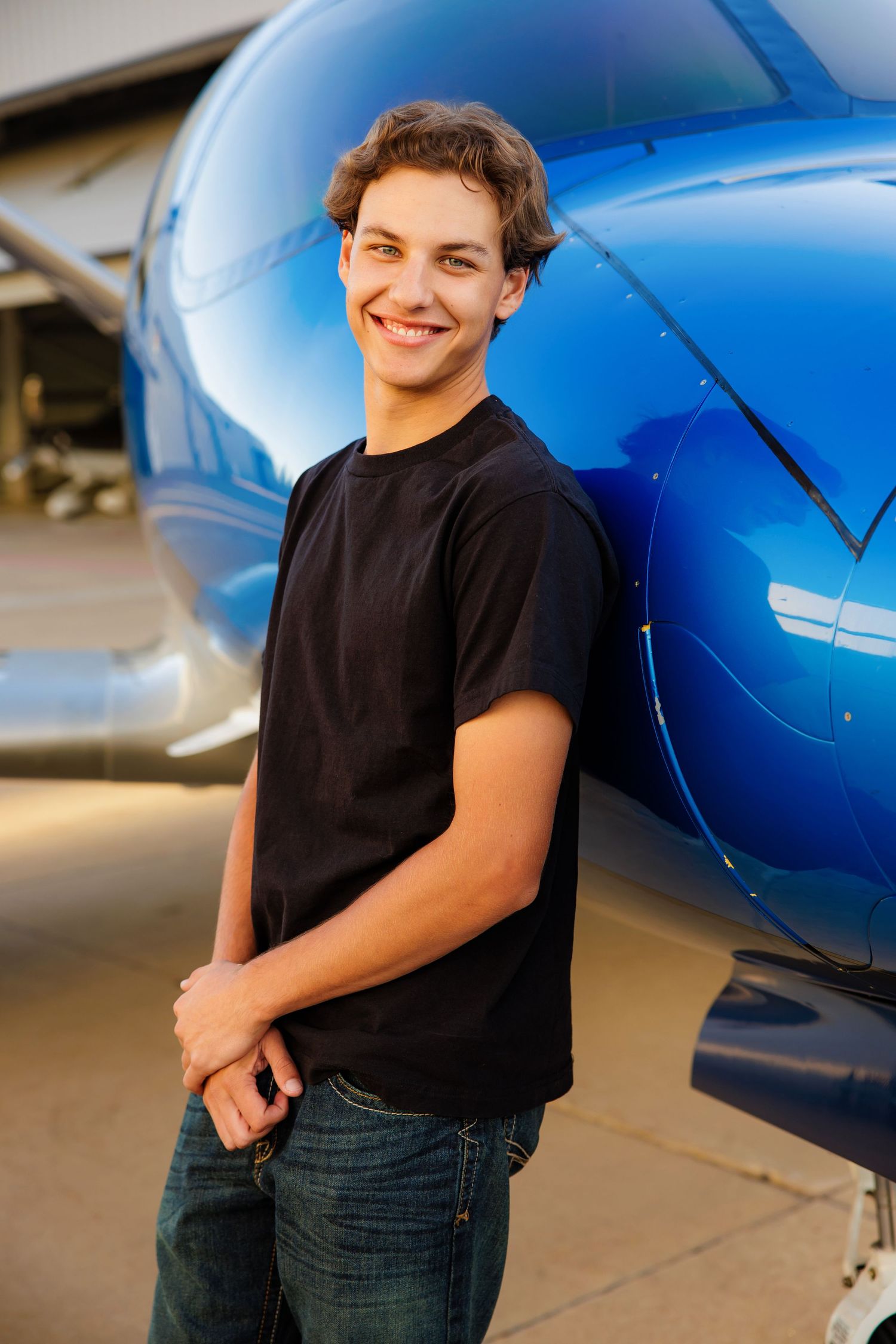 Smiling teenage boy in black t-shirt leans against a shiny blue aircraft outdoors.