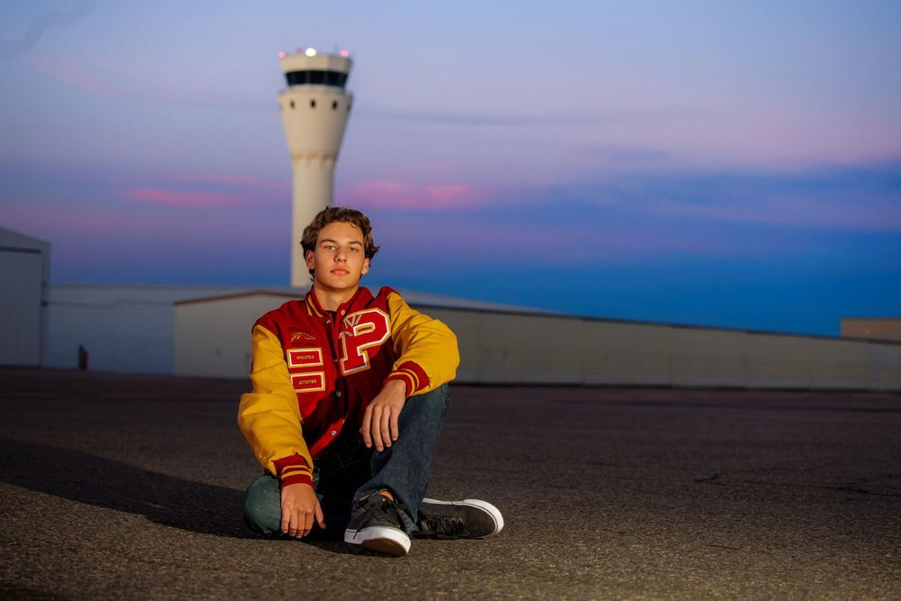 Teen in letterman jacket sitting on airport tarmac with air traffic control tower at dusk behind him. Centennial Colorado Senior Portrait