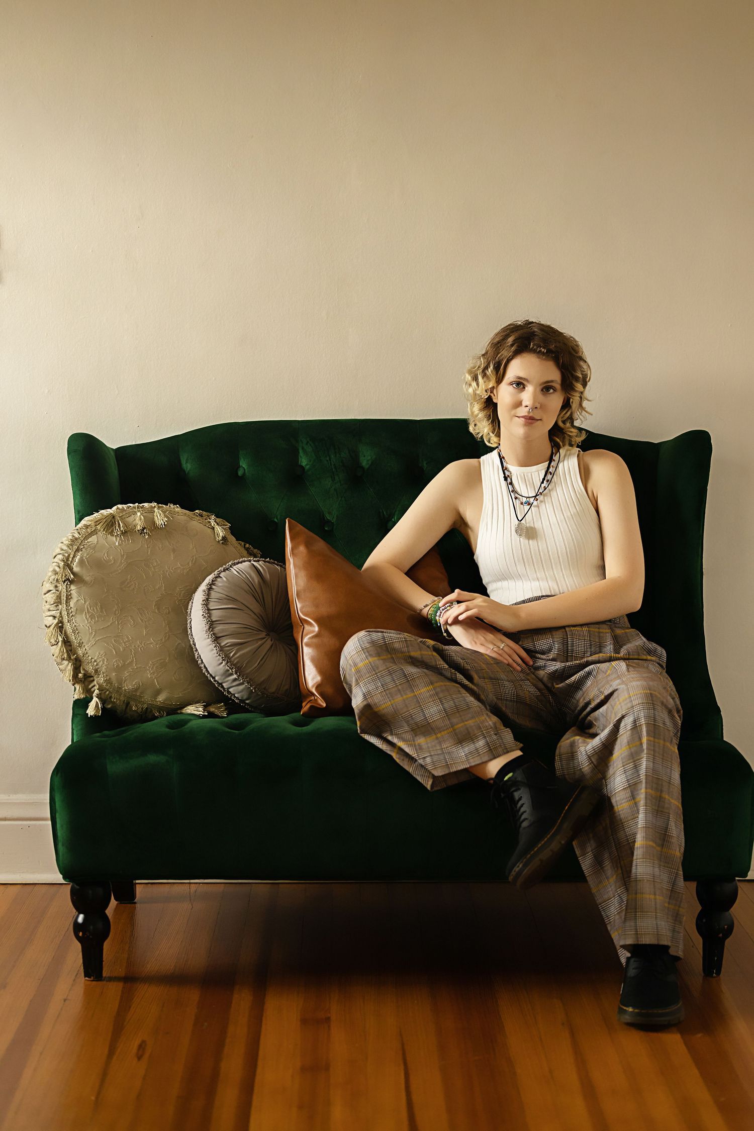 Androgynous teen in white tank and plaid pants sits relaxed on green tufted velvet sofa with pillows studio senior photo session
