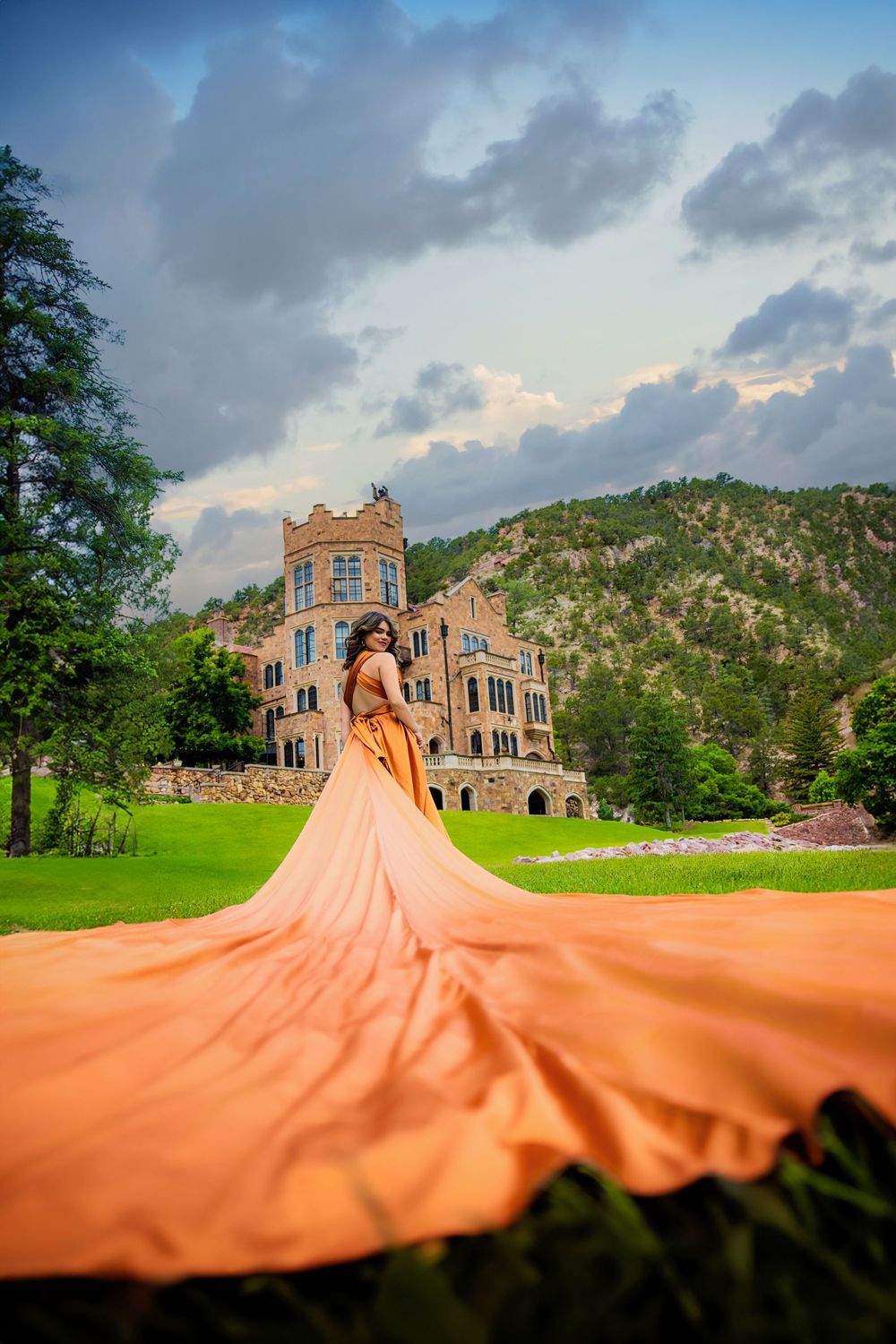 Woman in elegant orange gown with long trailing fabric posing before castle under dramatic cloudy sky for Parker Senior photos