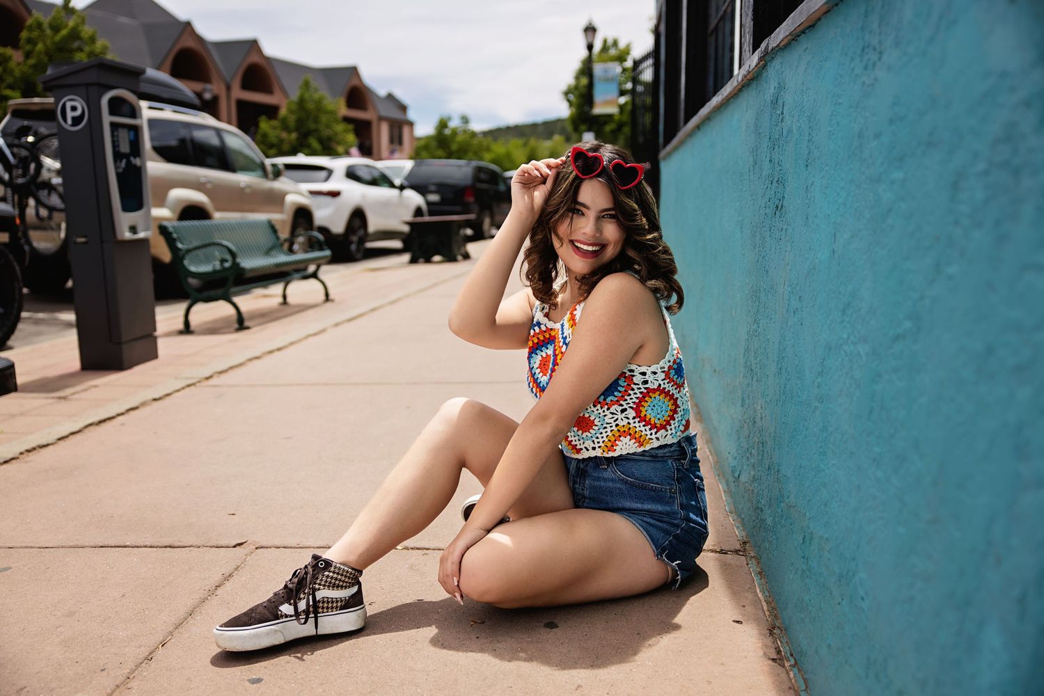Cheerful young woman in colorful crochet top and denim shorts sitting on urban sidewalk for Douglas County CO High School Senior pictures