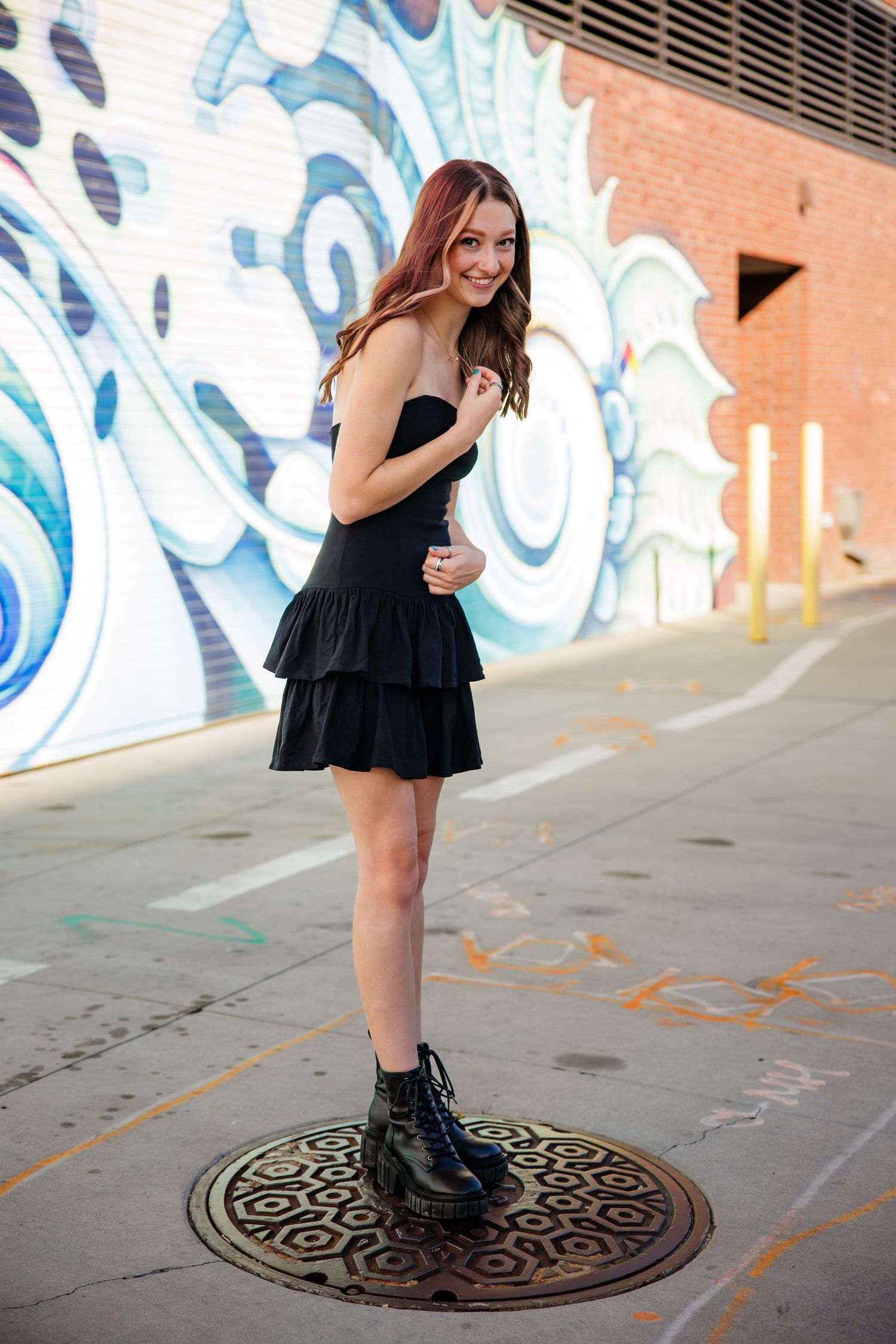 Smiling young woman in black mini dress and combat boots standing on urban street manhole cover for Aurora Colorado Senior pictures