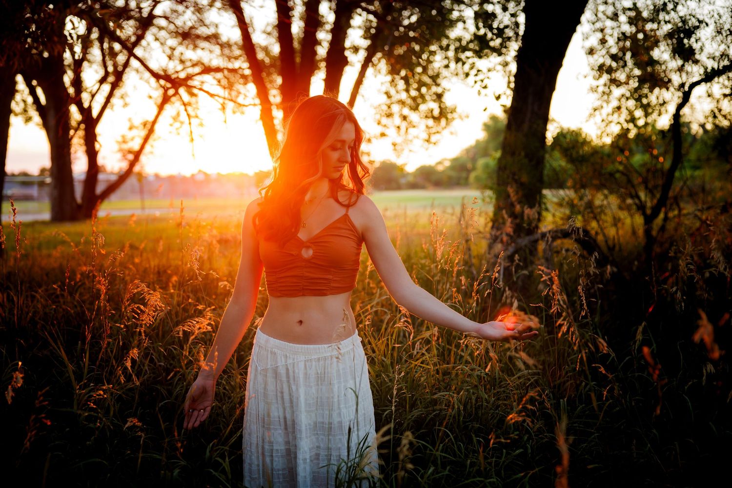 Woman in crop top and white skirt standing in tall grass field with vibrant orange sunset behind her for Parker CO Senior photos