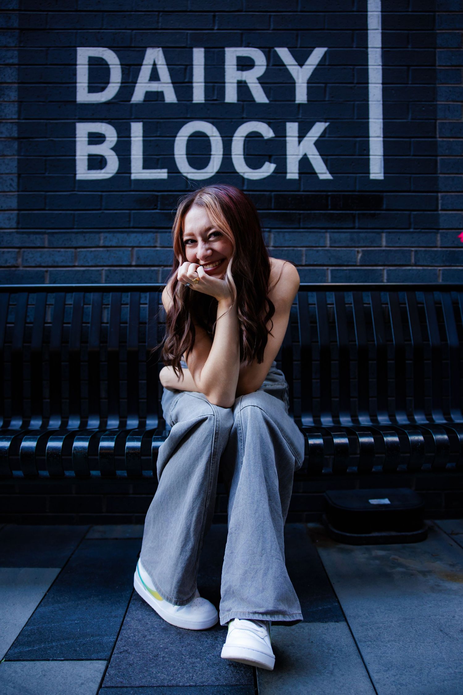 Young woman sitting on bench smiling in front of Dairy Block sign. Downtown Denver CO Senior photos