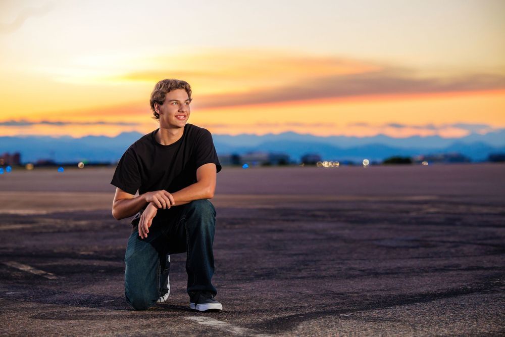 Teen in black outfit crouching on open tarmac at sunset with mountains and colorful sky in background. Parker Colorado Senior Portrait